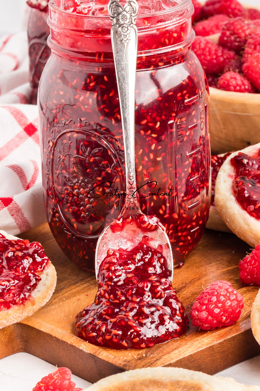 Jar of raspberry jam with a spoonful on a wooden board, surrounded by raspberries and bread.