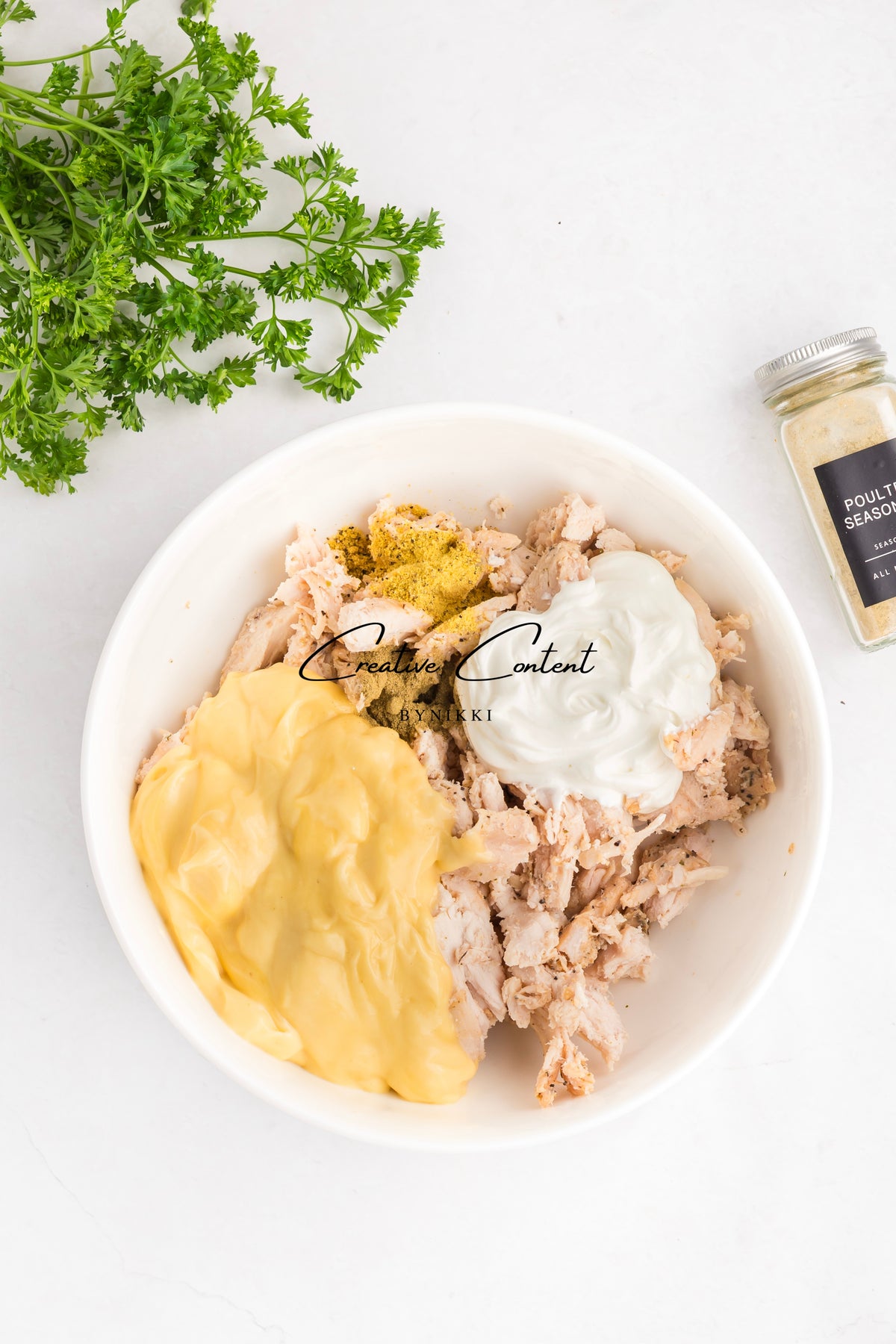 A white bowl containing Church Cookbook Chicken Casserole - Exclusive, featuring shredded chicken in a yellow creamy sauce topped with a dollop of sour cream and spices, set next to a bunch of parsley and a spice jar.