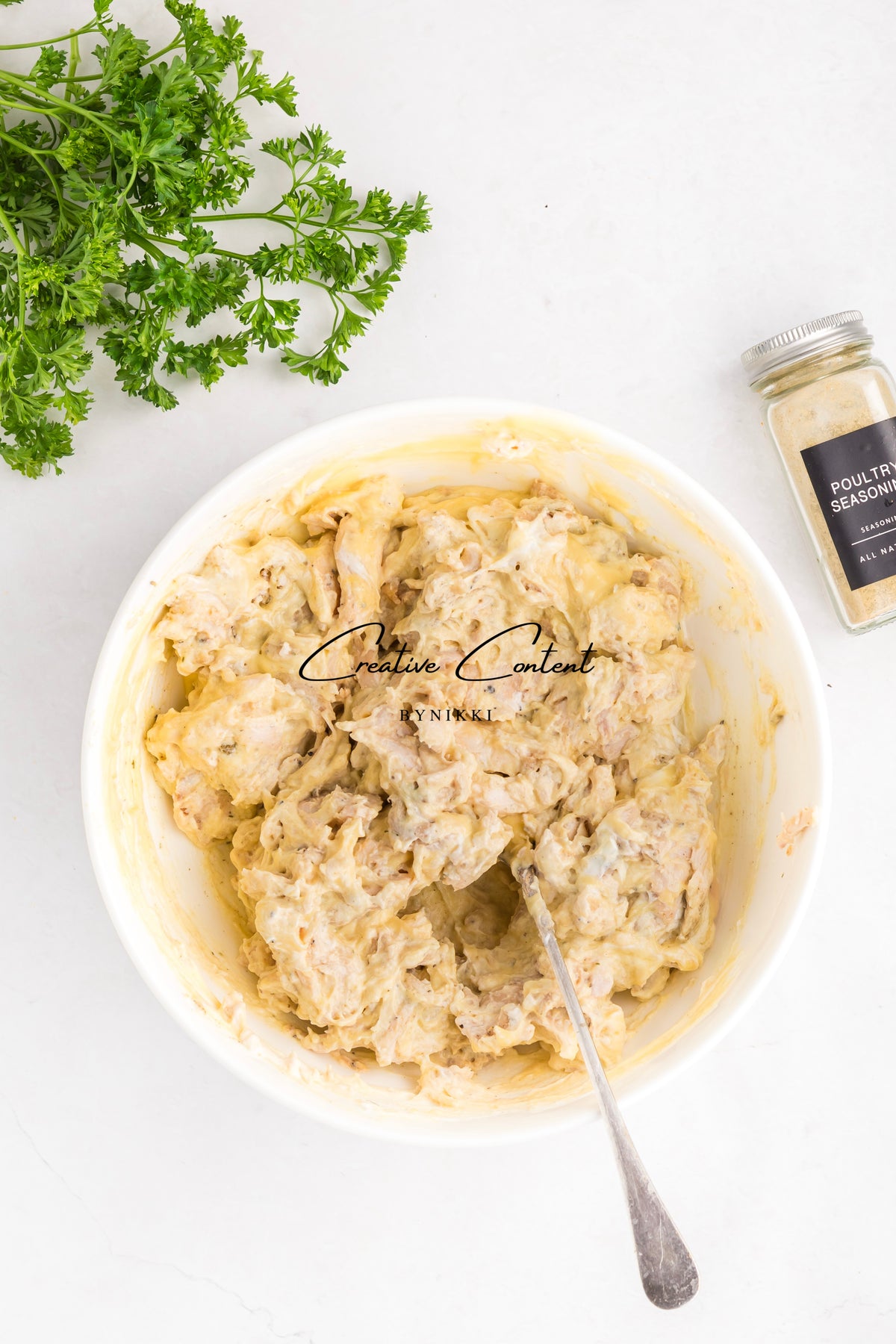 A bowl of creamy Church Cookbook Chicken Casserole - Exclusive mixture with a spoon, accompanied by fresh parsley and a jar of poultry seasoning.