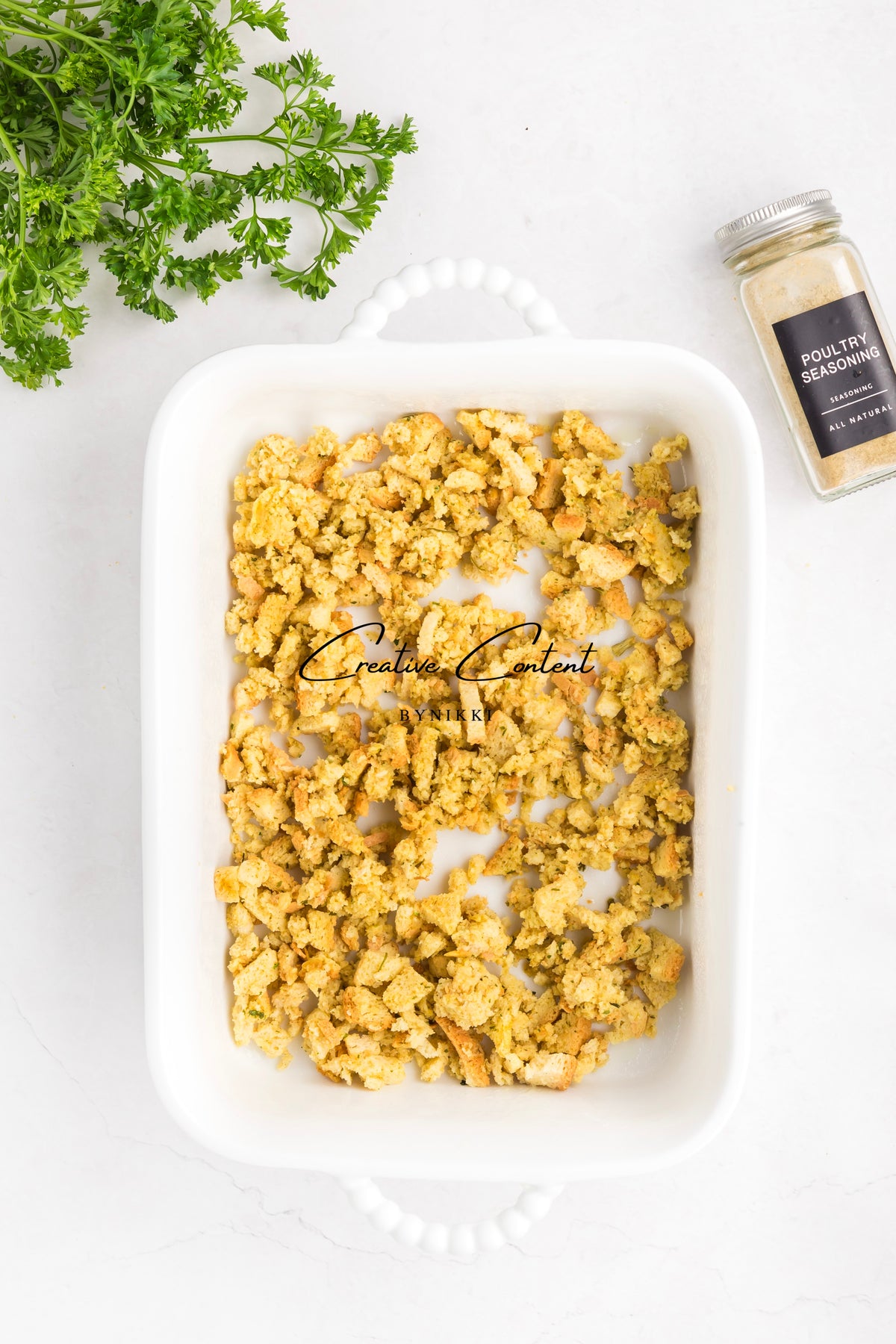 A white rectangular casserole dish filled with seasoned breadcrumbs for a Church Cookbook Chicken Casserole - Exclusive, set next to a sprig of parsley and a jar of poultry seasoning.