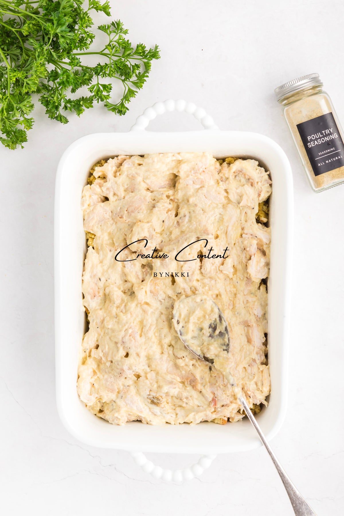 A white casserole dish filled with a creamy Church Cookbook Chicken Casserole - Exclusive, served with a spoon, fresh parsley, and a jar of poultry seasoning nearby.