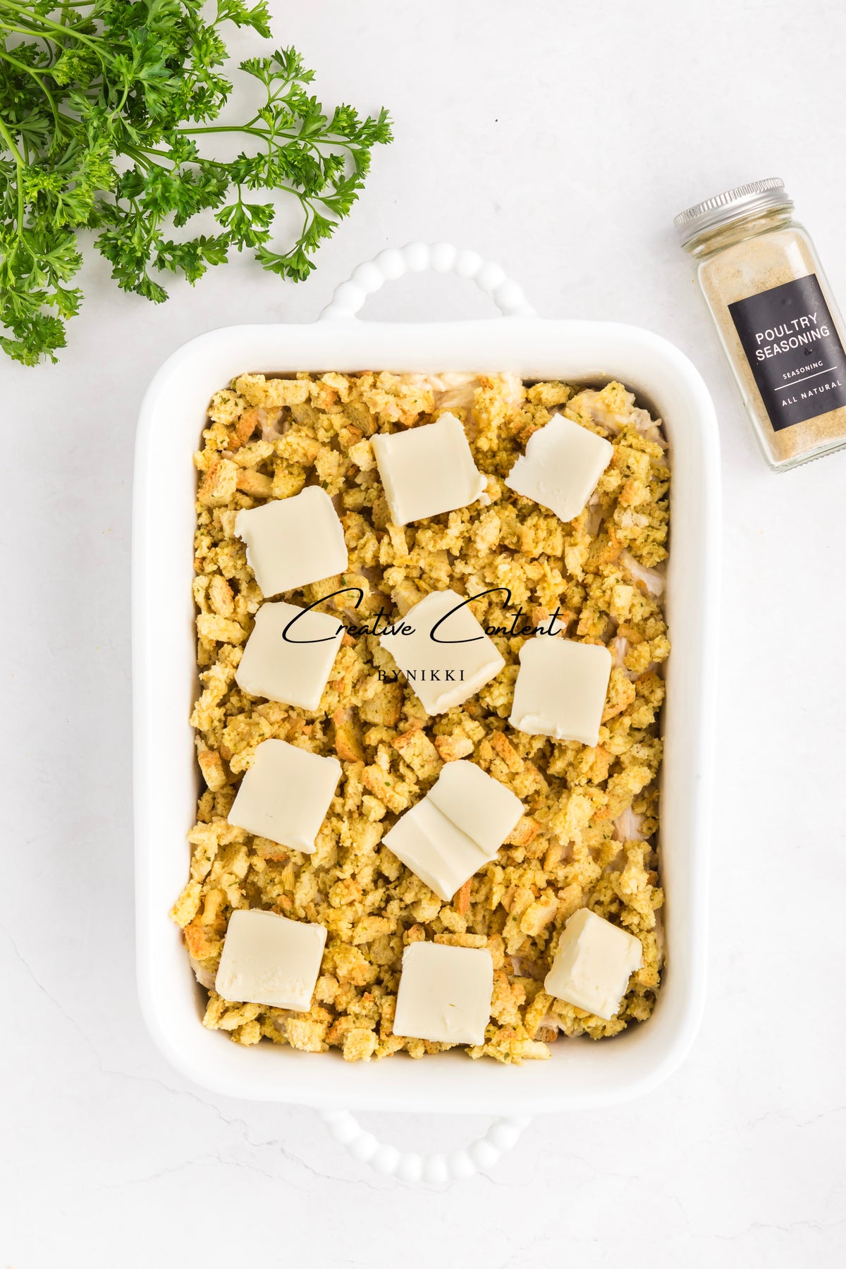 A white baking dish filled with Church Cookbook Chicken Casserole - Exclusive, topped with breadcrumbs and pats of butter, with parsley and a jar of poultry seasoning nearby.