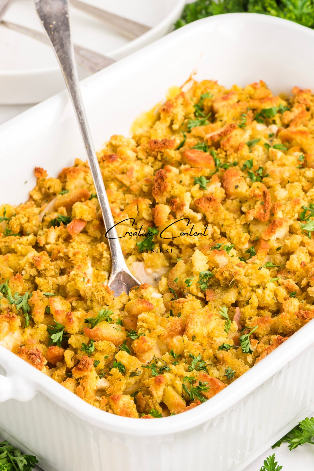A white baking dish filled with a Church Cookbook Chicken Casserole - Exclusive topped with golden brown crumbs and sprinkled with fresh parsley, with a serving spoon resting inside.