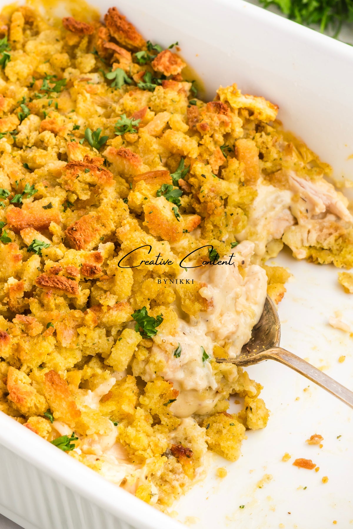 A white baking dish filled with a Church Cookbook Chicken Casserole - Exclusive, topped with crispy breadcrumbs and fresh herbs, with a spoon serving a portion.