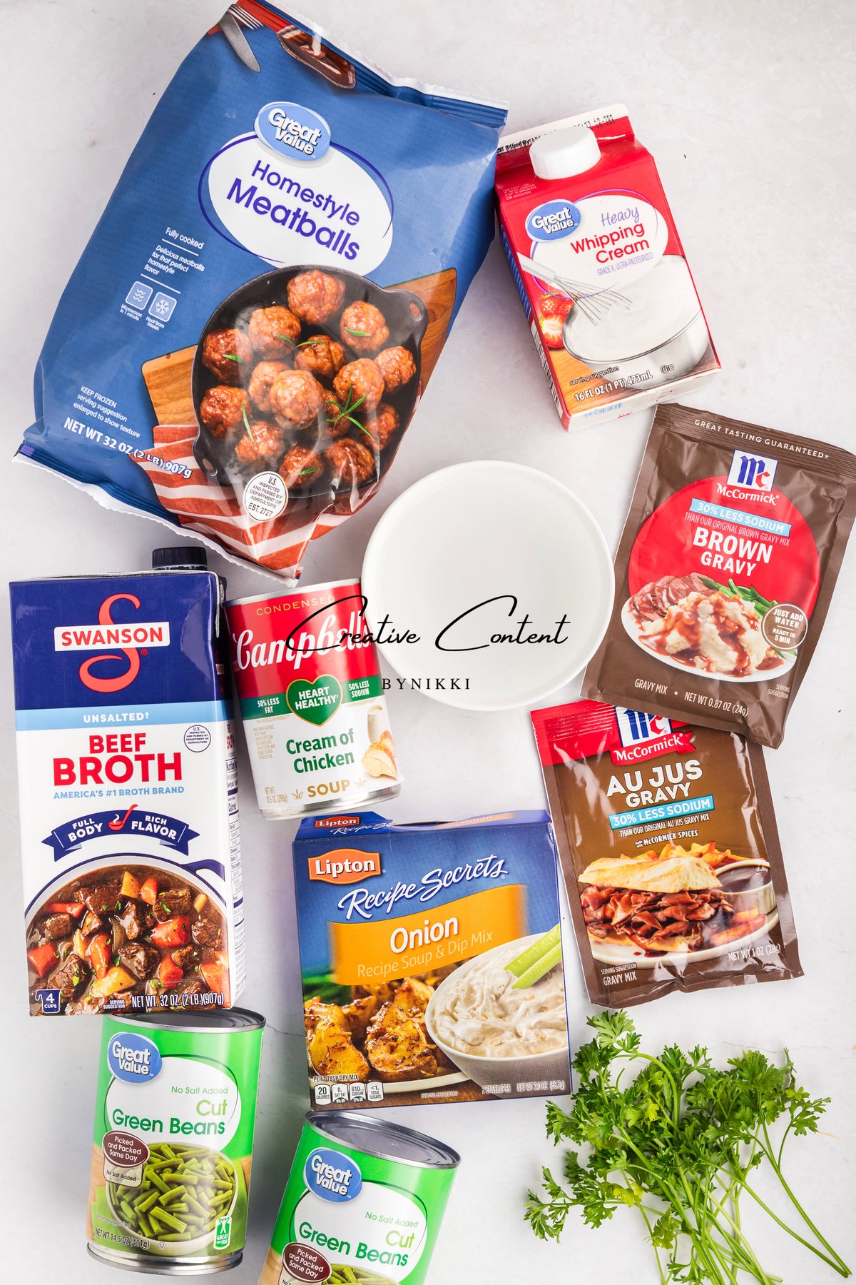 Ingredients for Crock Pot Smothered Meatballs and Green Beans - Exclusive, including frozen meatballs, beef broth, heavy cream, cream of chicken soup, green beans, onion soup mix, gravy packets, and fresh parsley surrounding an empty white bowl.