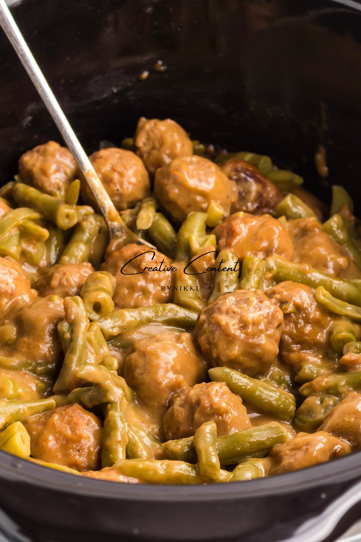 A slow cooker filled with Crock Pot Smothered Meatballs and Green Beans - Exclusive, consisting of meatballs and green beans coated in a brown gravy, with a serving spoon resting in the center.