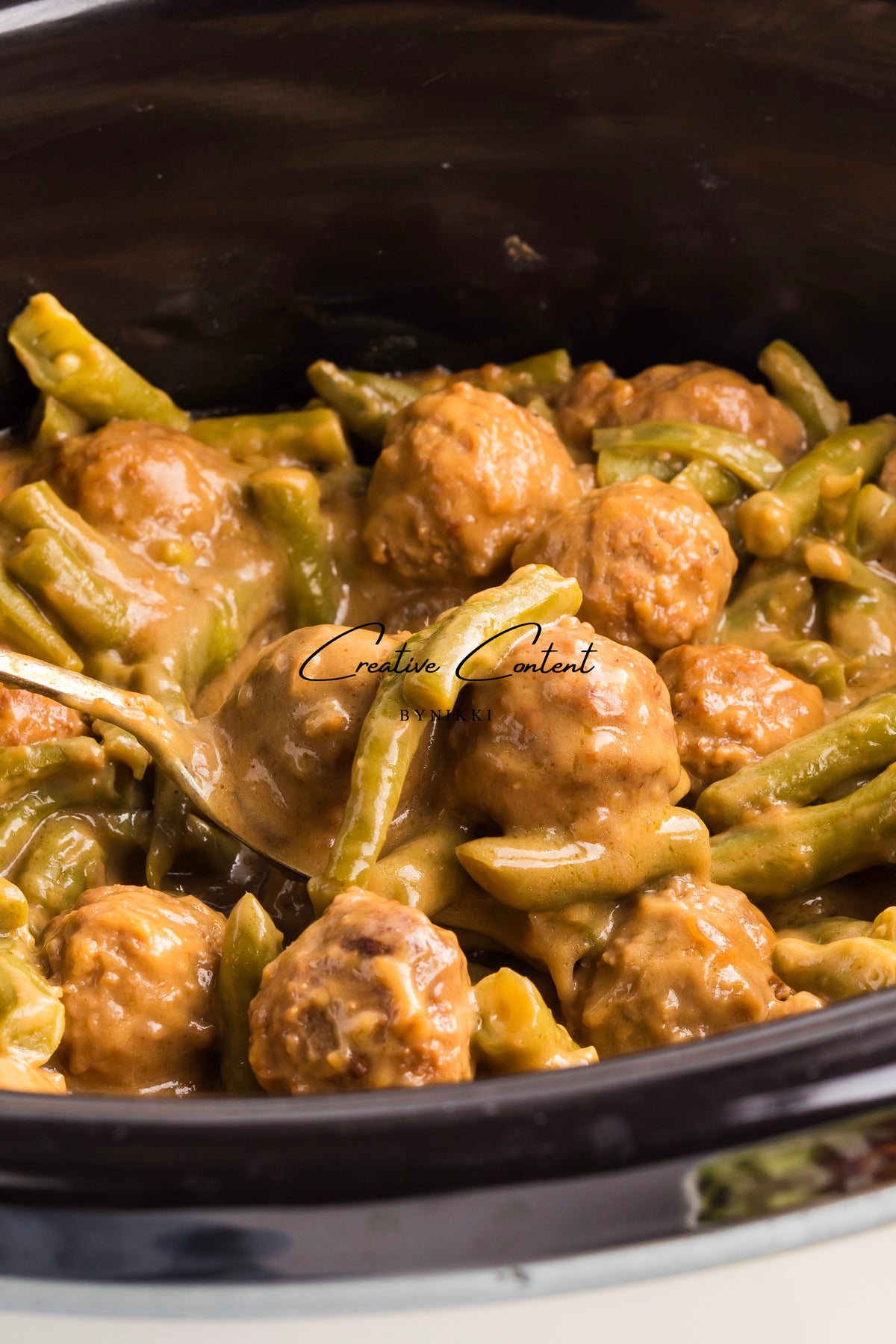 A serving spoon lifts Crock Pot Smothered Meatballs and Green Beans - Exclusive from a slow cooker, showing meatballs and green beans coated in a creamy brown sauce.
