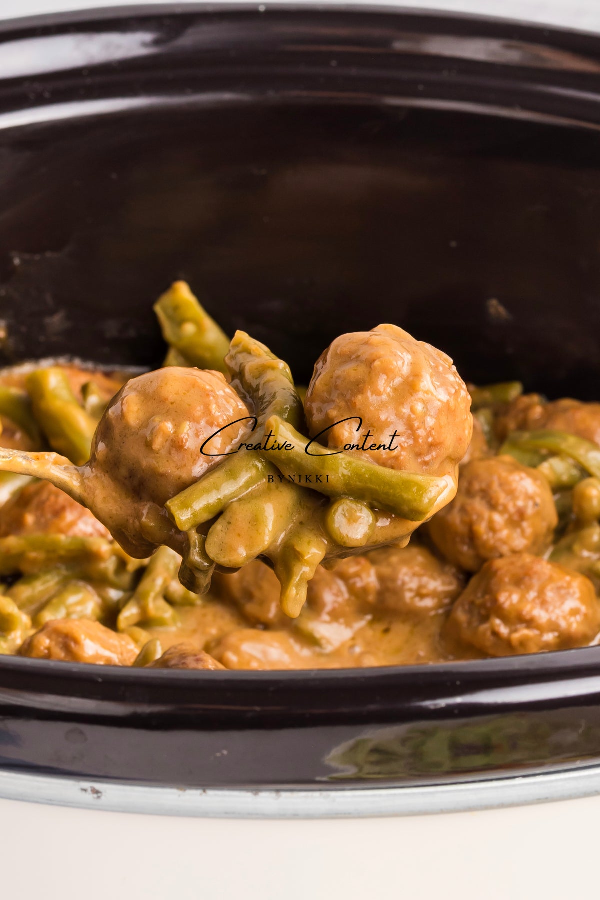A serving spoon lifts Crock Pot Smothered Meatballs and Green Beans - Exclusive in a creamy sauce from a slow cooker.