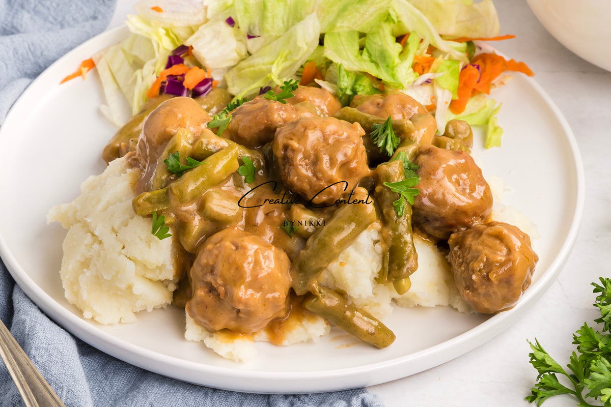 Crock Pot Smothered Meatballs and Green Beans - Exclusive served over a scoop of mashed potatoes with a side salad on a white plate.