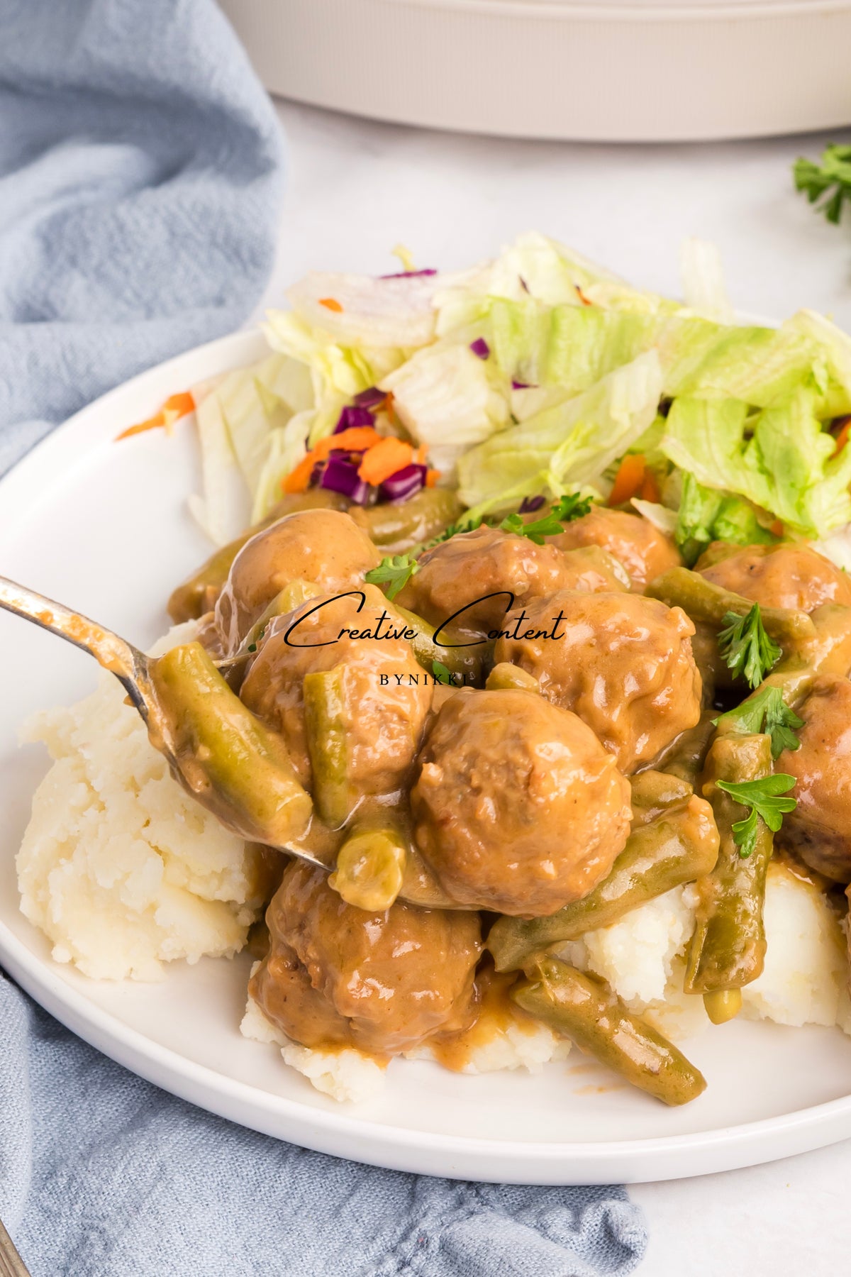 A spoonful of Crock Pot Smothered Meatballs and Green Beans - Exclusive served over mashed potatoes with a side of salad on a white plate.