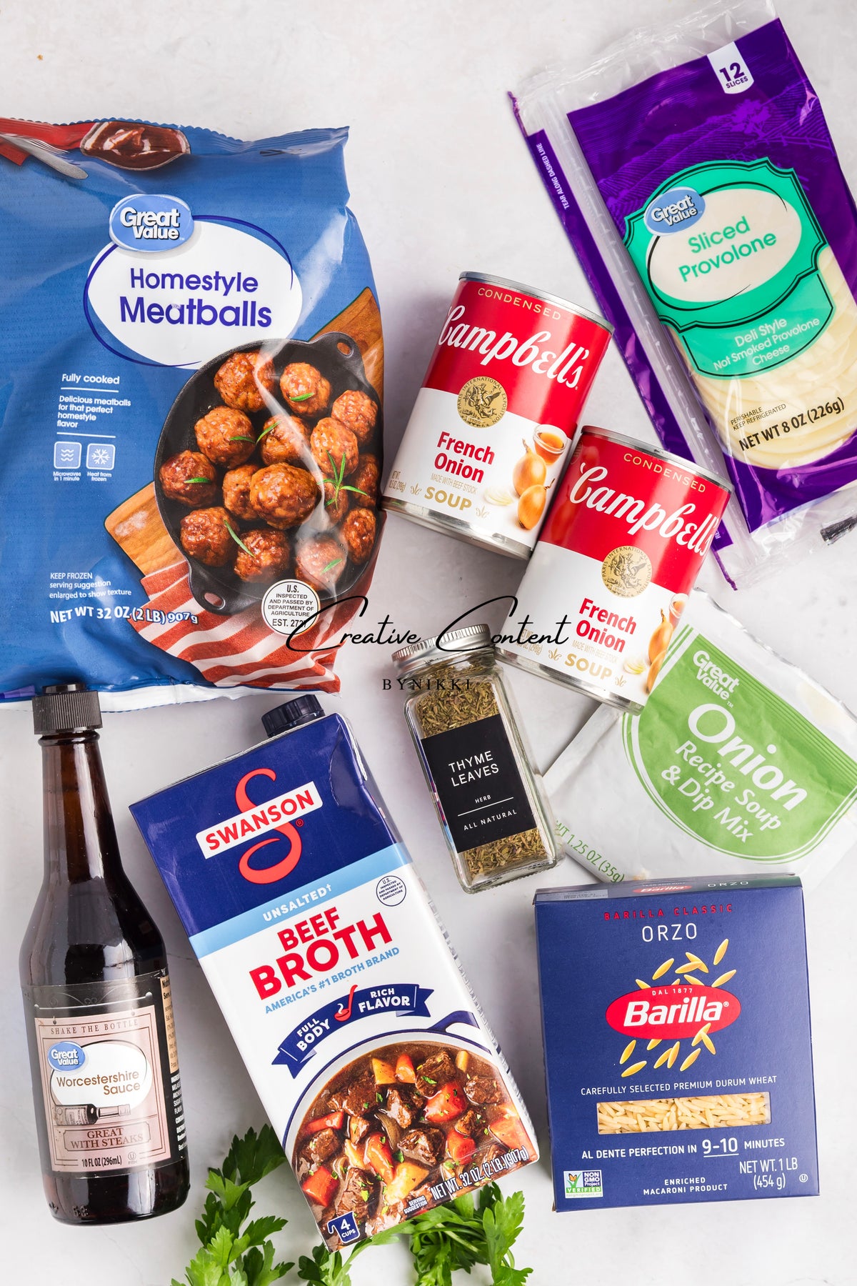 Ingredients for a French Onion Meatball Orzo Bake - Exclusive, including frozen meatballs, two cans of French onion soup, provolone, orzo, beef broth, Worcestershire sauce, onion soup mix, and dried thyme arranged on a white surface.