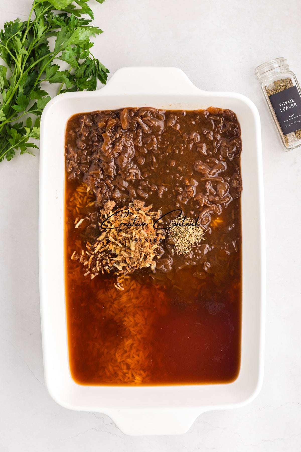 A rectangular white baking dish holding the ingredients for a French Onion Meatball Orzo Bake - Exclusive, including broth, orzo, caramelized onions, and dried herbs, with parsley and a jar of thyme nearby.