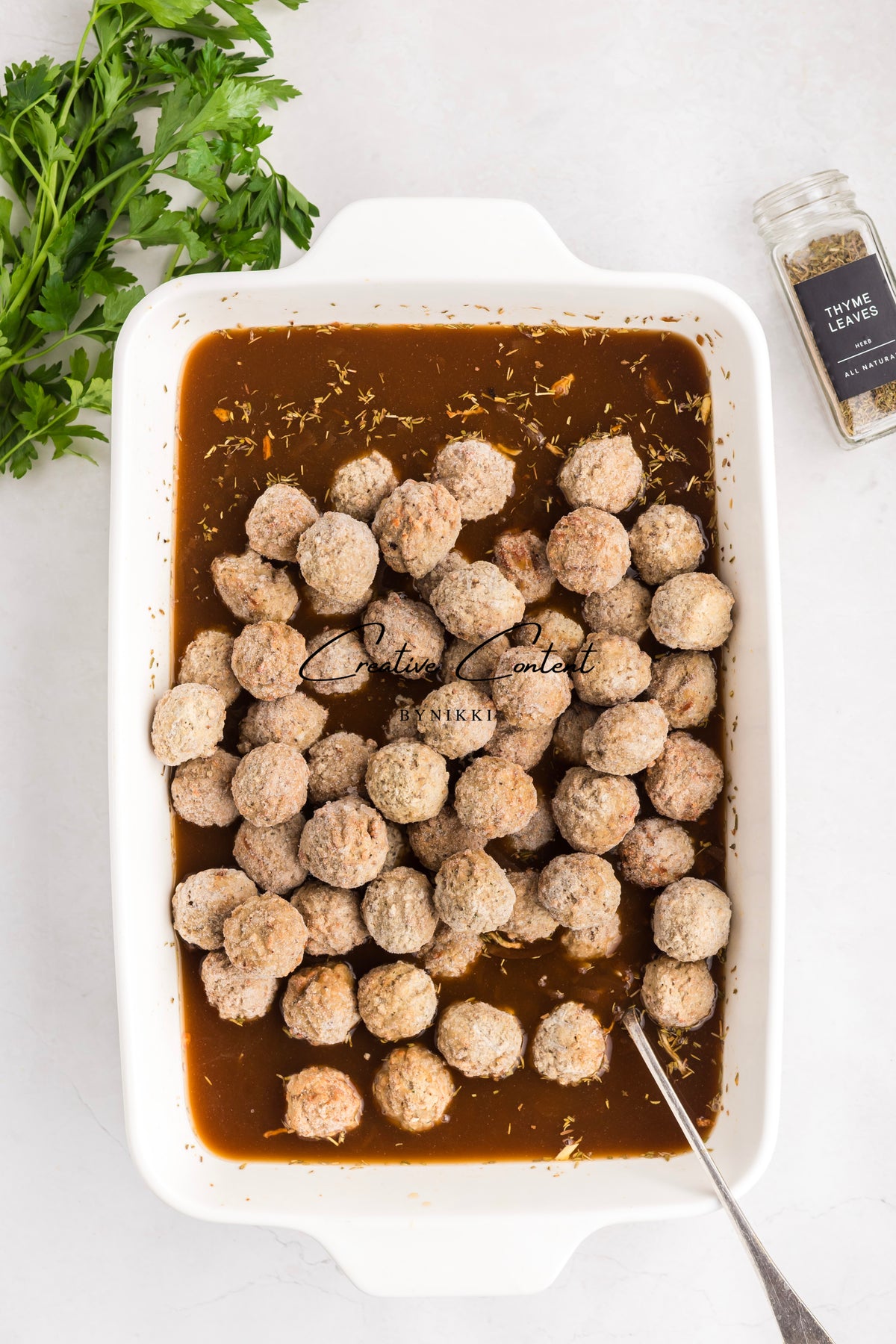 Meatballs in brown sauce in a white baking dish with parsley and thyme leaves