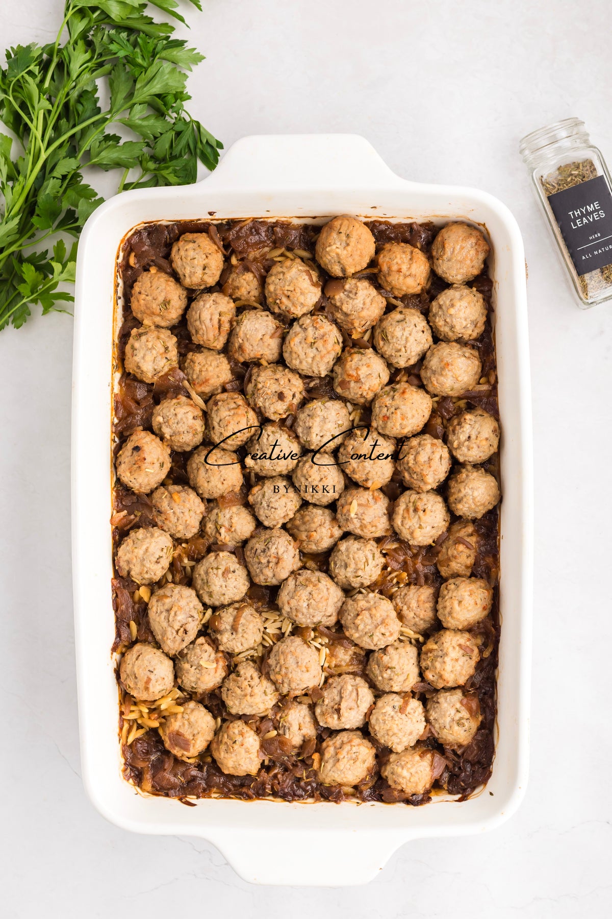 A rectangular casserole dish of French Onion Meatball Orzo Bake - Exclusive, topped with meatballs and a sprig of parsley, with a jar of thyme leaves on the side.