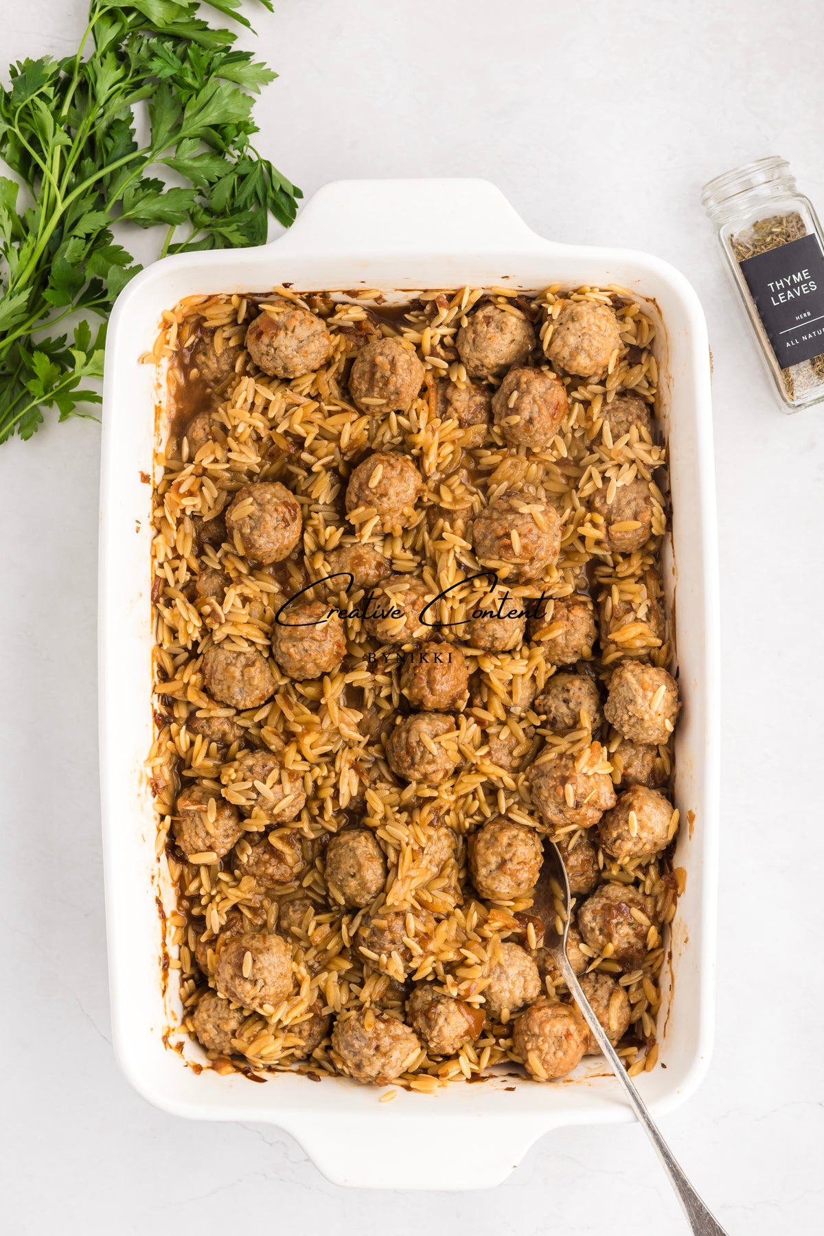A white baking dish filled with a French Onion Meatball Orzo Bake - Exclusive, accompanied by a serving spoon, fresh parsley, and a jar of thyme.