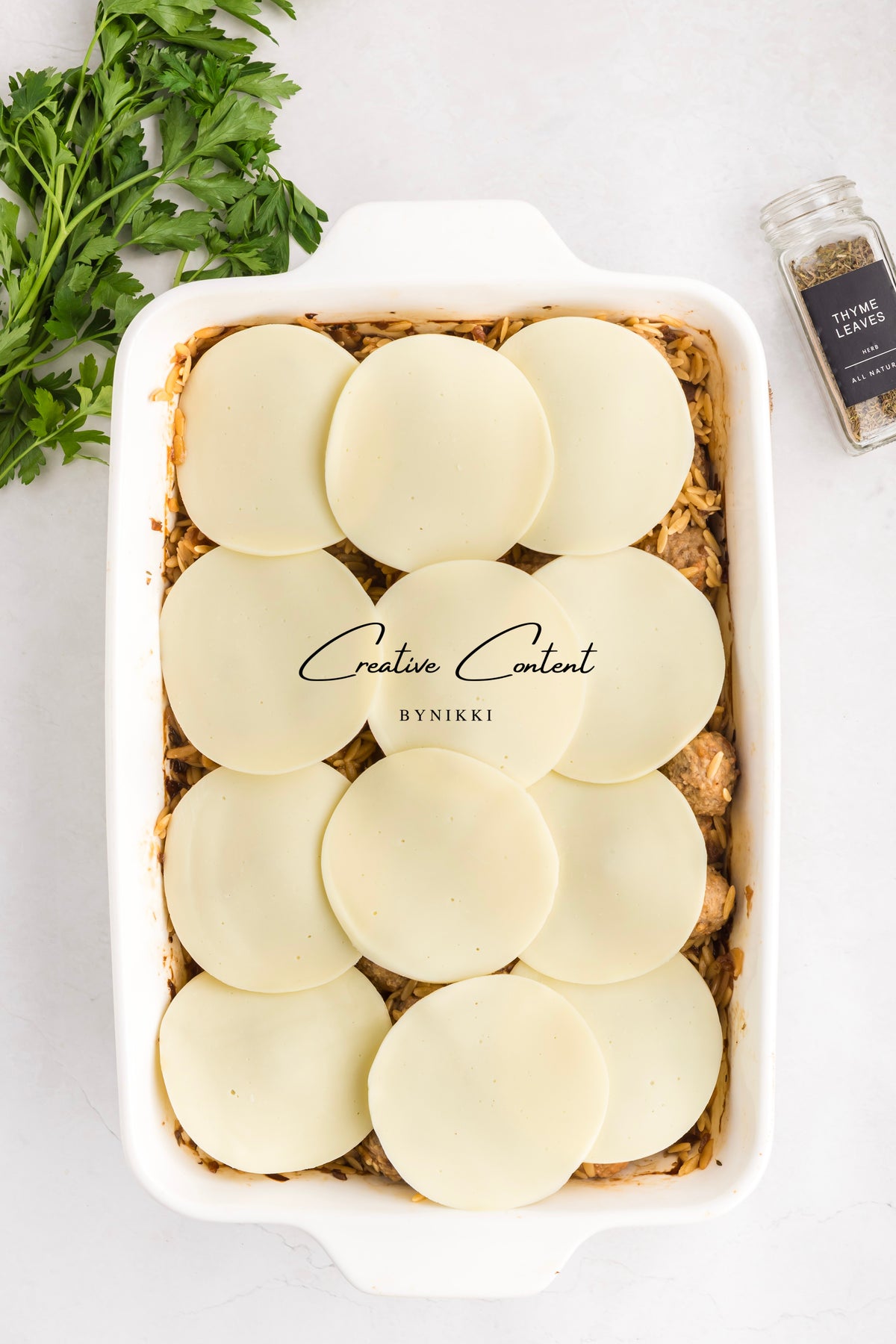 A white baking dish filled with a French Onion Meatball Orzo Bake - Exclusive, topped with round slices of cheese, next to parsley and a jar of thyme.