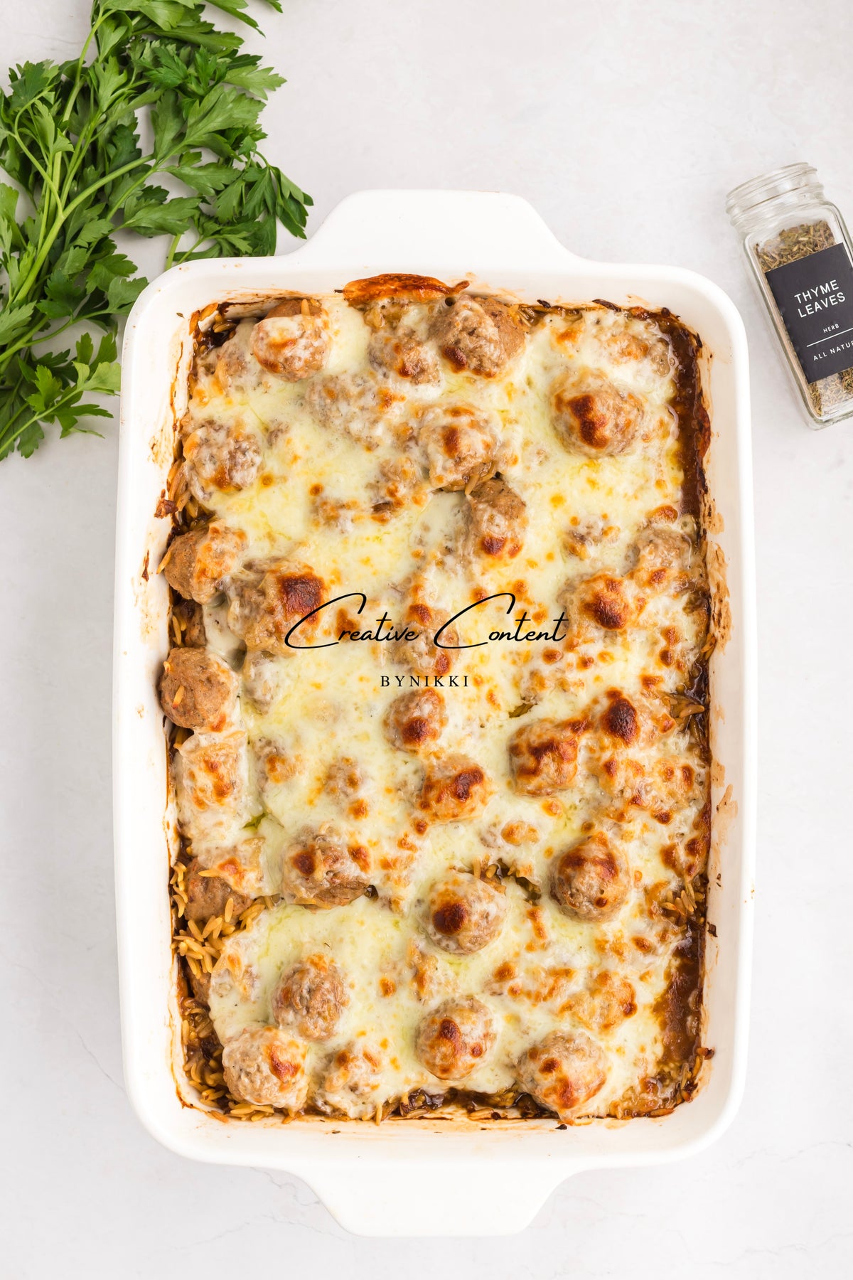 A white rectangular baking dish filled with a French Onion Meatball Orzo Bake - Exclusive topped with melted cheese, with fresh parsley and a jar of thyme nearby.