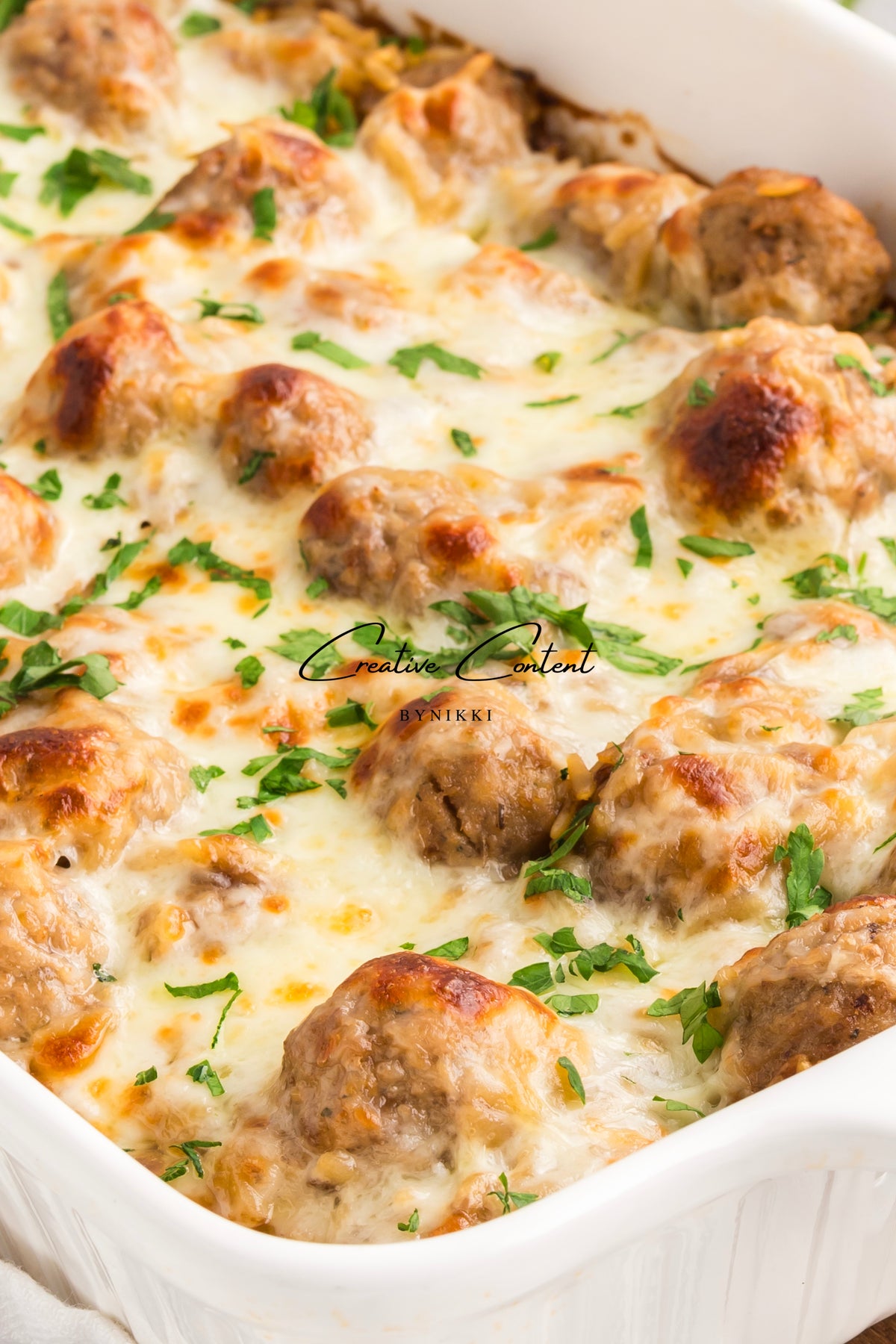 A white baking dish filled with a French Onion Meatball Orzo Bake, featuring meatballs topped with melted cheese and fresh parsley.