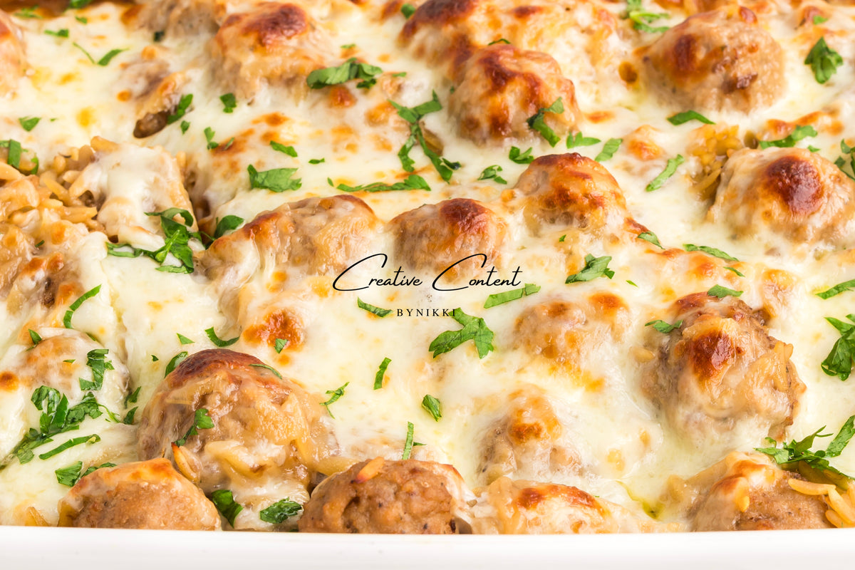 A French Onion Meatball Orzo Bake - Exclusive, consisting of meatballs and orzo pasta covered in melted cheese and garnished with herbs.