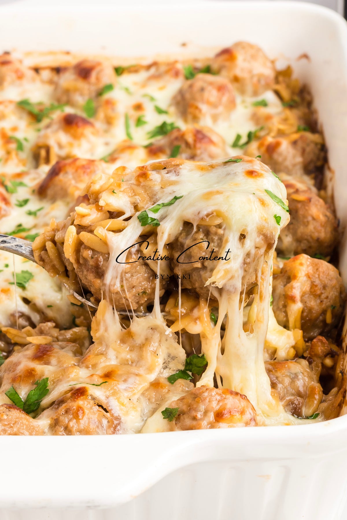 A serving spoon lifts meatballs and orzo from a French Onion Meatball Orzo Bake - Exclusive, with long strands of melted cheese stretching from the serving.