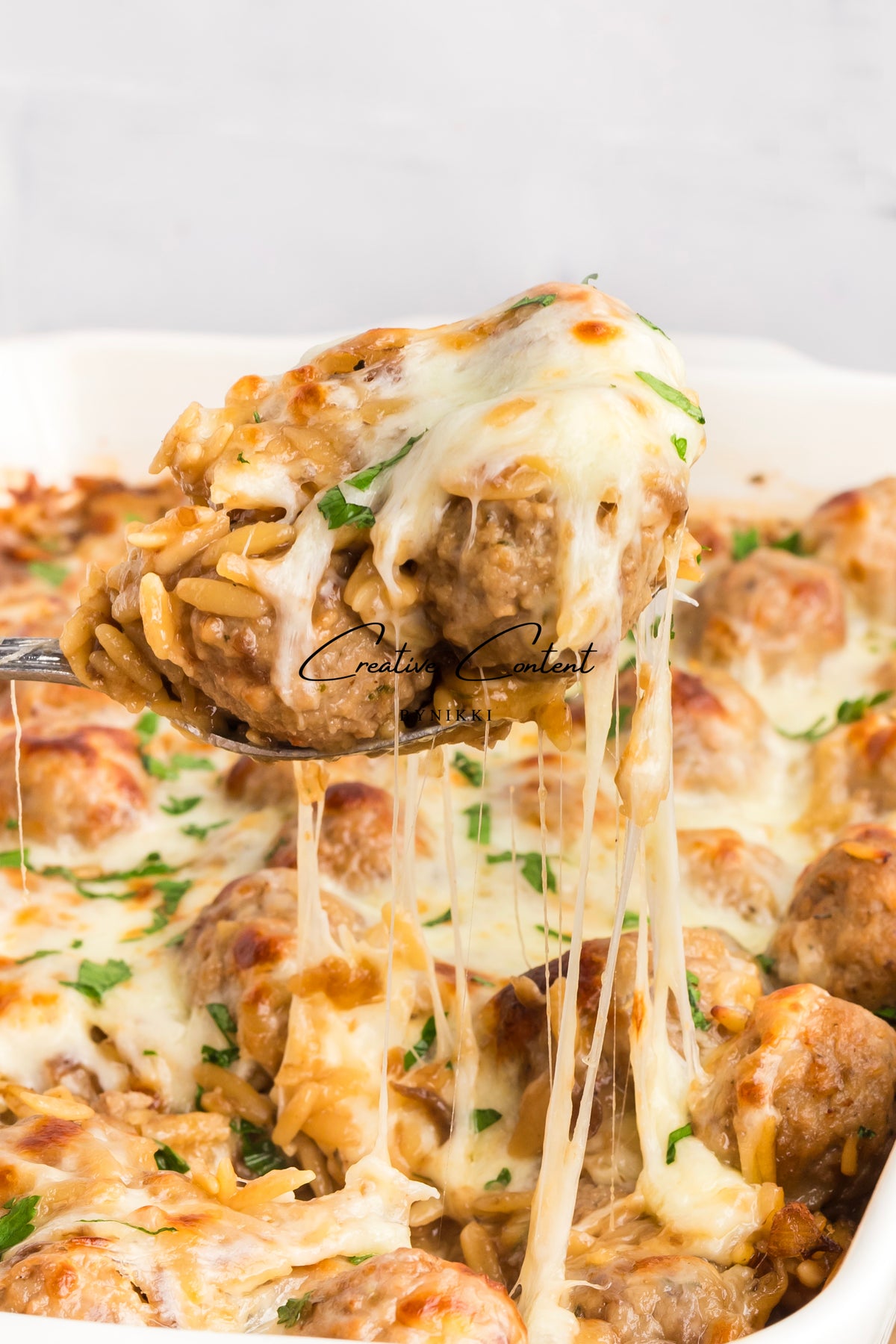 Spoon lifting French Onion Meatball Orzo Bake with melted cheese and herbs