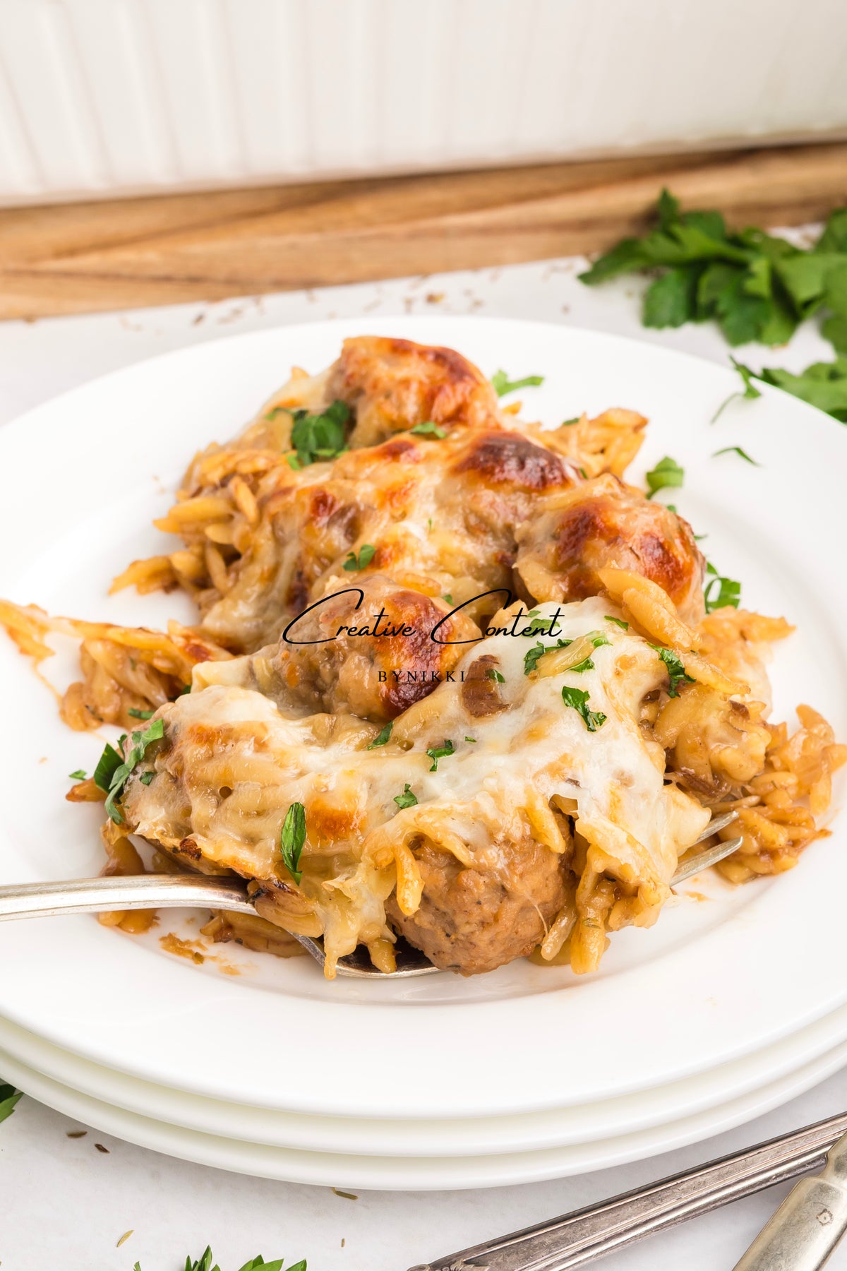 A serving of French Onion Meatball Orzo Bake - Exclusive, topped with melted cheese and fresh herbs, on a white plate with a fork.