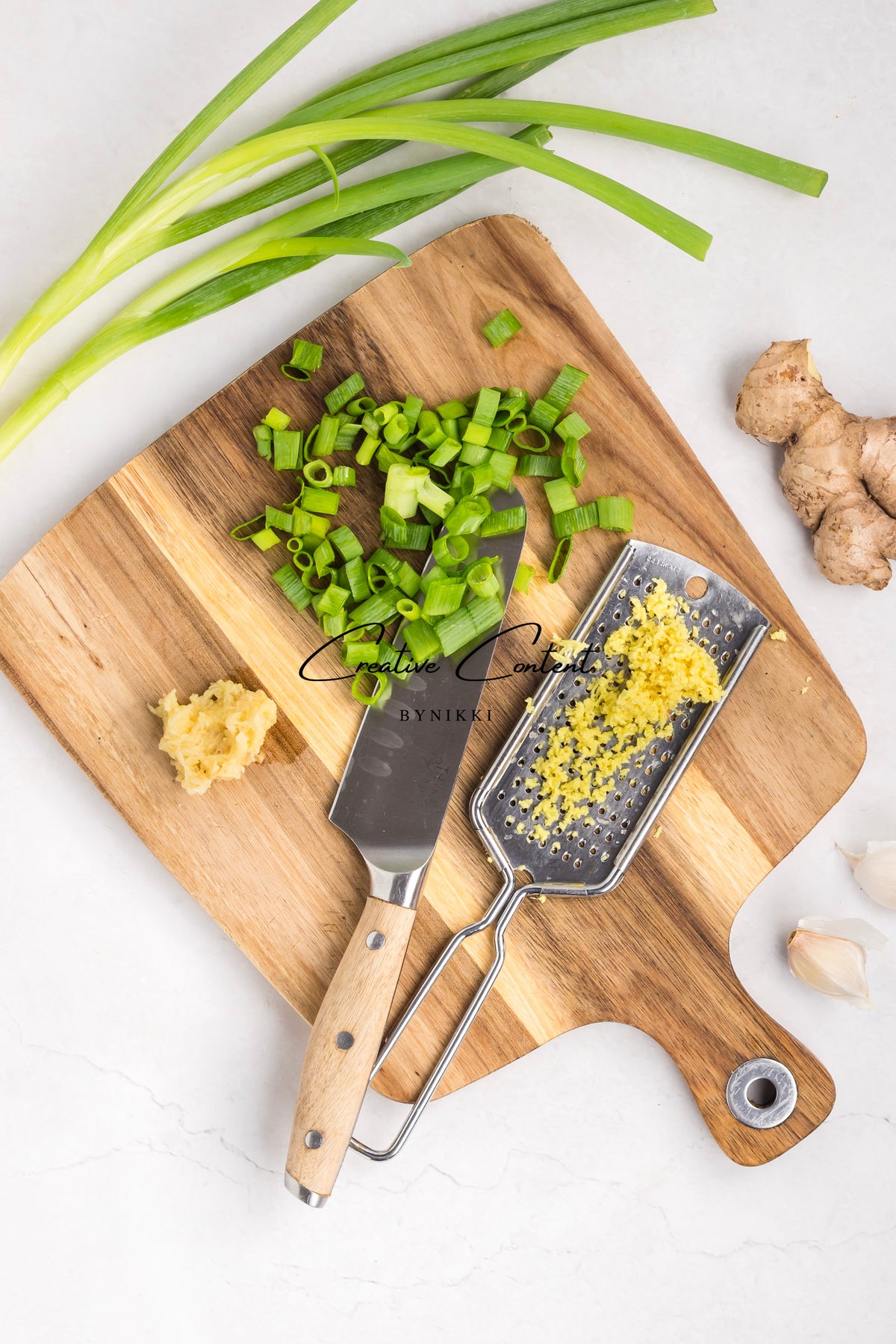 A wooden cutting board with chopped green onions, a chef’s knife, a ginger grater, ginger root, and garlic cloves for a Korean Beef Bulgogi - Exclusive recipe.