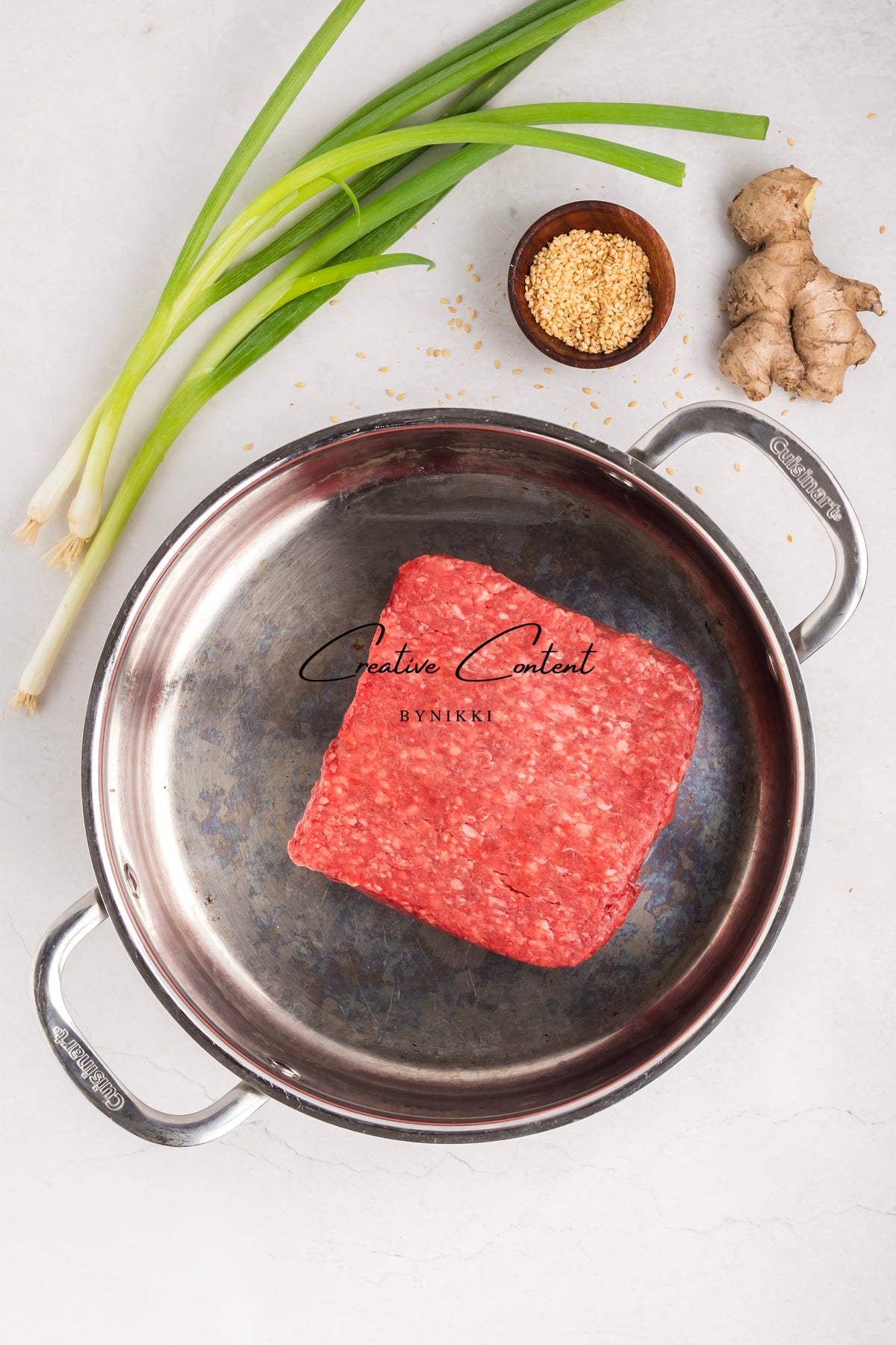 A square block of raw ground beef for Korean Beef Bulgogi - Exclusive sits in a stainless steel skillet, surrounded by green onions, a knob of ginger, and a small bowl of sesame seeds.