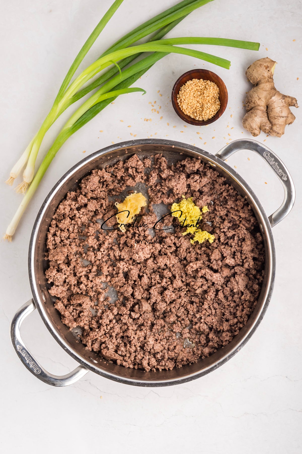 A skillet of browned ground meat for a Korean Beef Bulgogi - Exclusive recipe, featuring two dollops of minced aromatics, green onions, a bowl of sesame seeds, and ginger root.