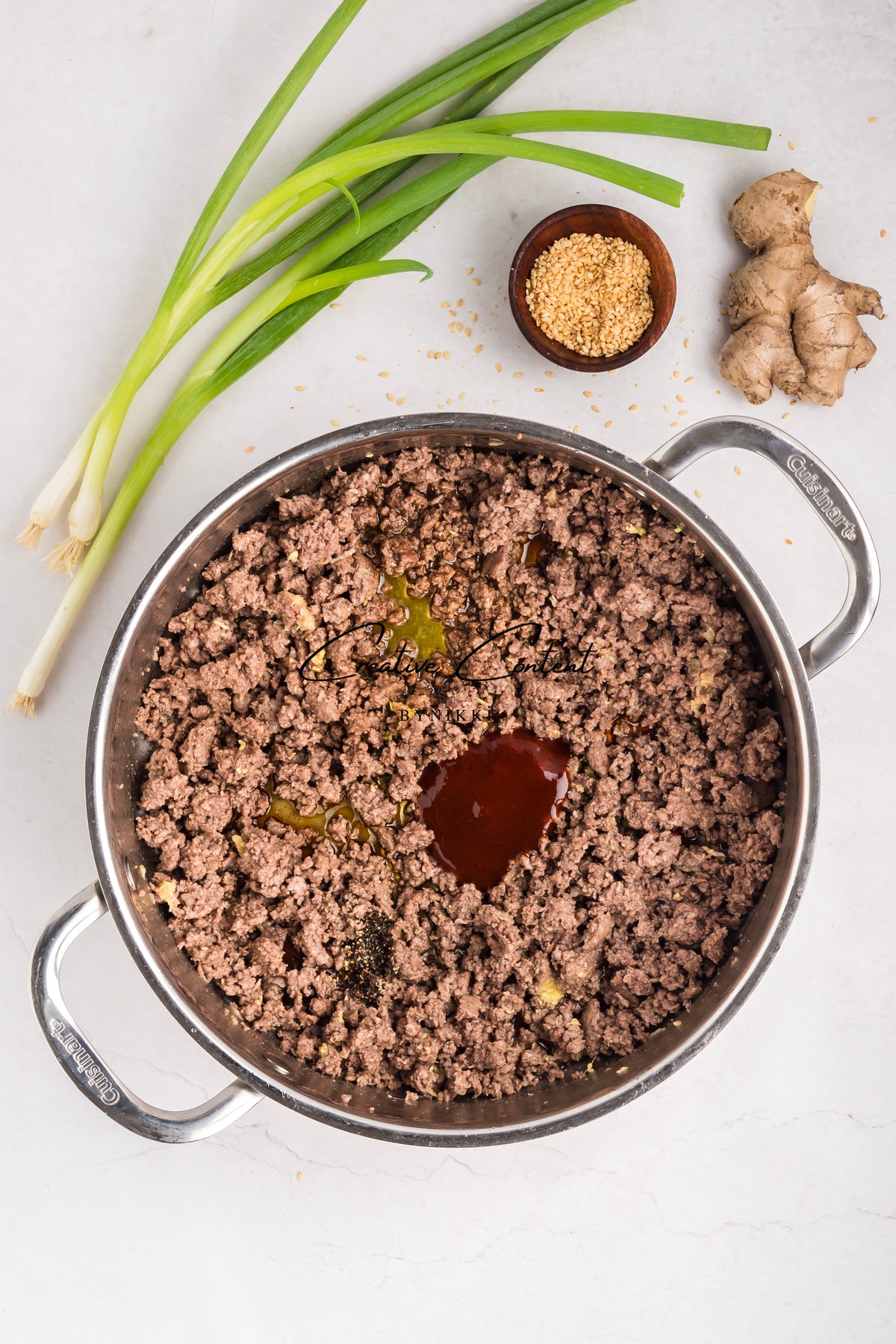 A skillet of Korean Beef Bulgogi - Exclusive featuring ground beef in gochujang sauce, accompanied by green onions, a ginger root, and a small bowl of sesame seeds.