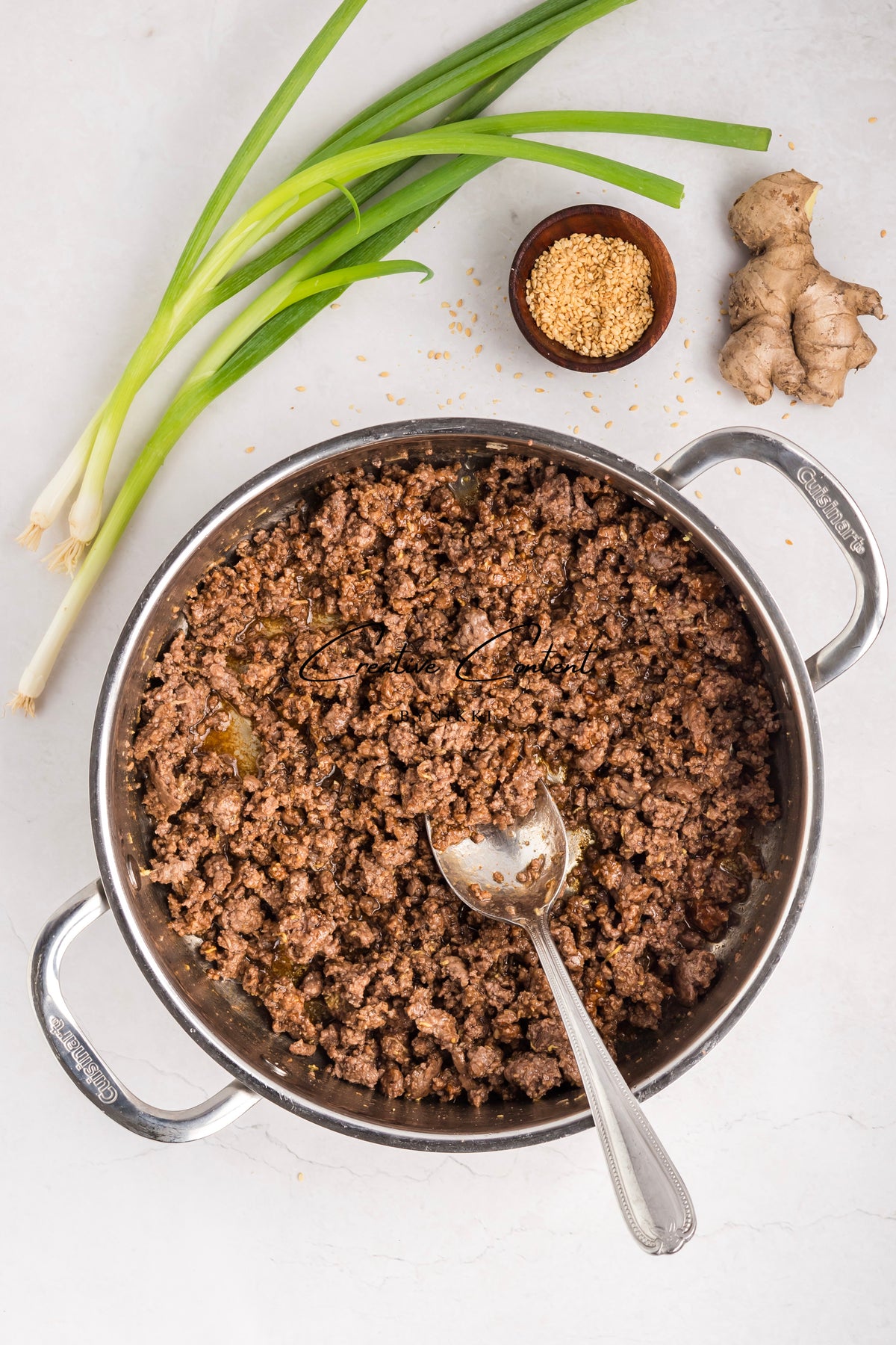 A stainless steel pan filled with Korean Beef Bulgogi - Exclusive for a rice bowl and a serving spoon, surrounded by green onions, a small bowl of sesame seeds, and a piece of ginger.