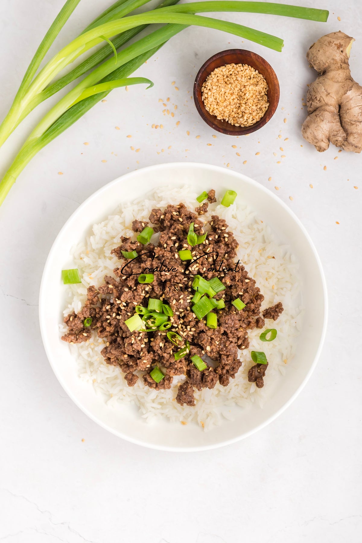 A Korean Beef Bulgogi - Exclusive bowl of gochujang-sauced ground beef served over white rice, topped with sliced green onions and sesame seeds, surrounded by fresh ginger and a small dish of sesame seeds.