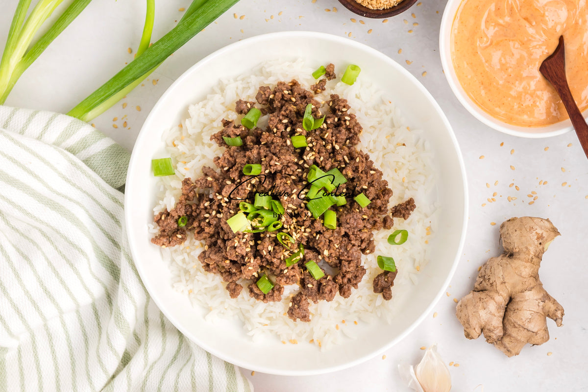 Korean Beef Bulgogi - Exclusive ground beef rice bowl topped with sesame seeds and chopped green onions, served with a side of gochujang sauce and fresh ginger.