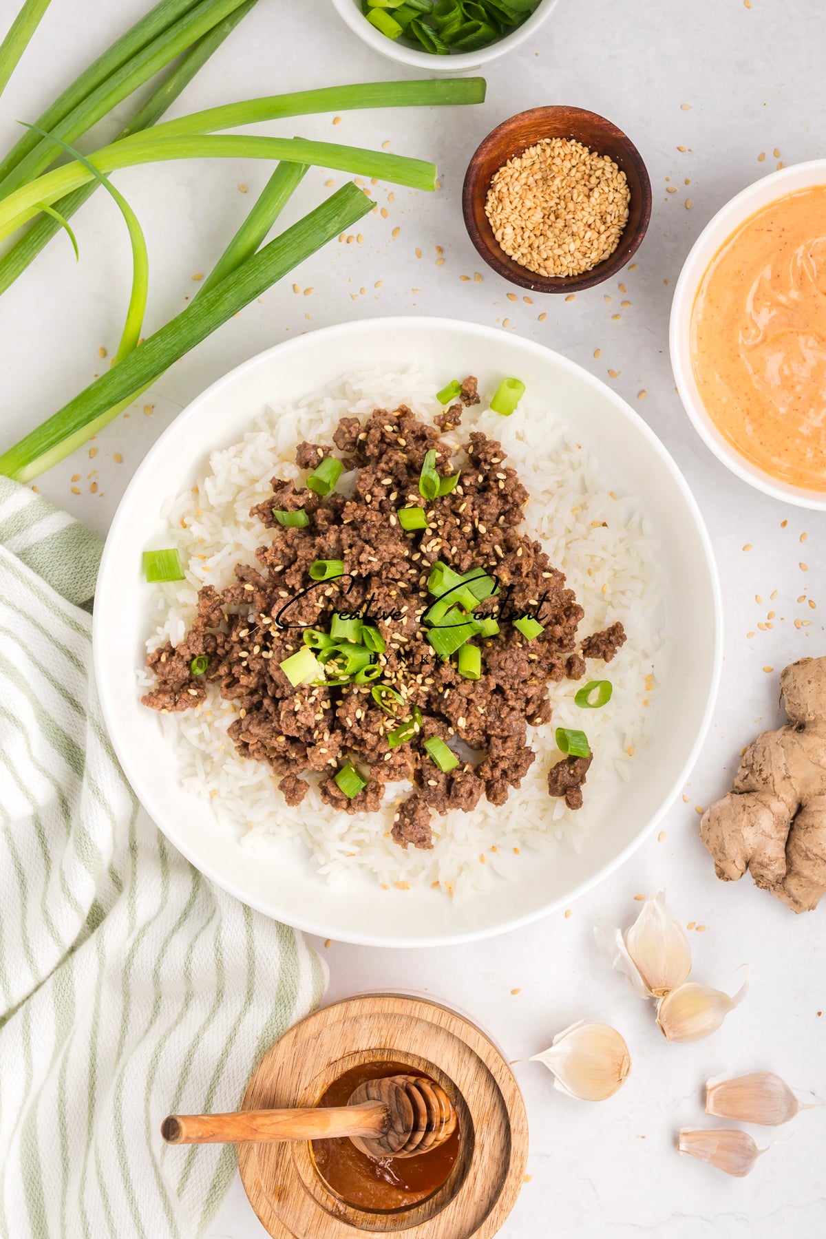 A bowl of Korean Beef Bulgogi - Exclusive featuring ground beef and gochujang sauce over white rice, garnished with green onions and sesame seeds, surrounded by scallions, extra sauce, garlic, ginger, and honey.