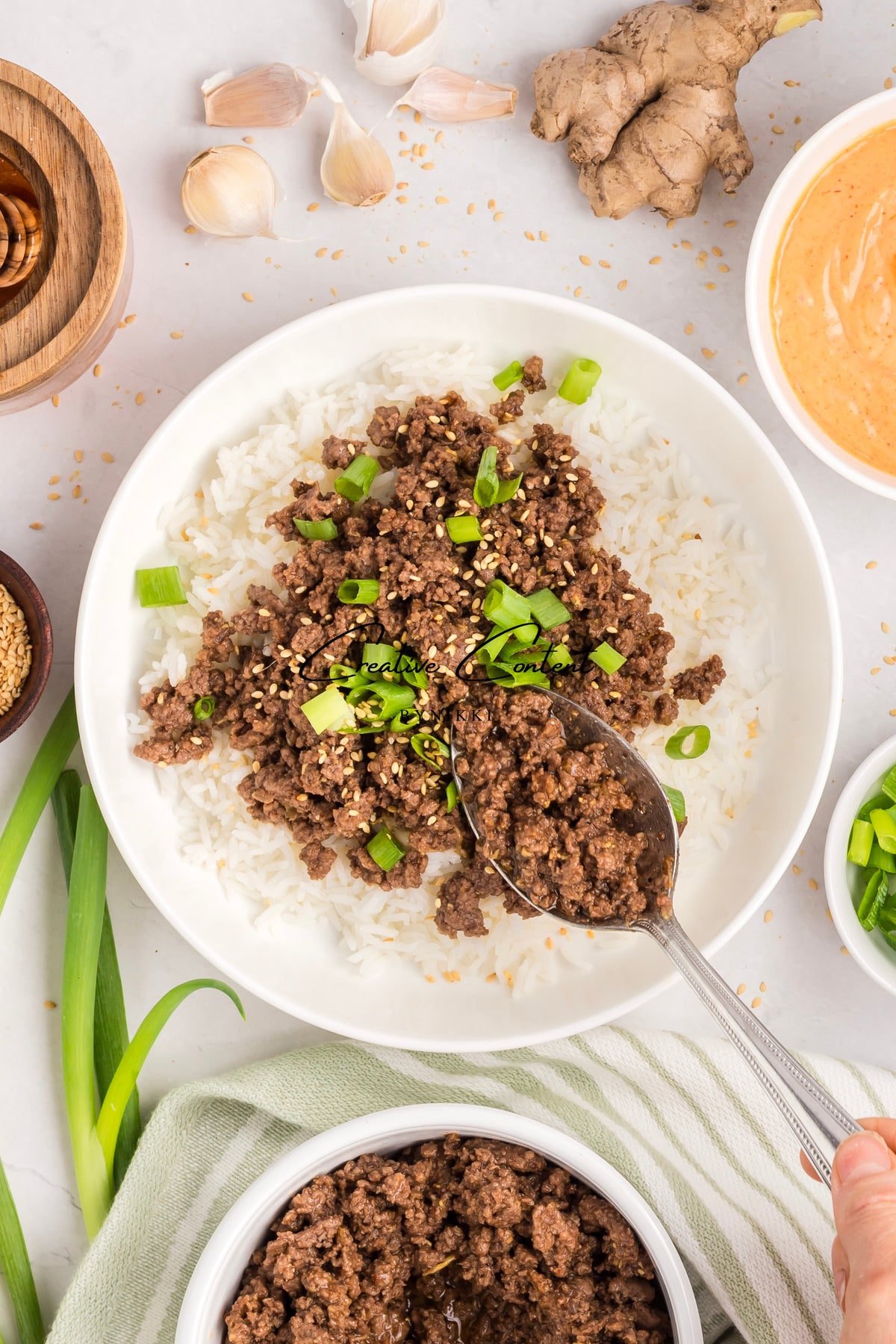 A spoon lifts a portion of Korean Beef Bulgogi - Exclusive, topped with sesame seeds and green onions, from a white bowl with gochujang sauce and fresh aromatics nearby.
