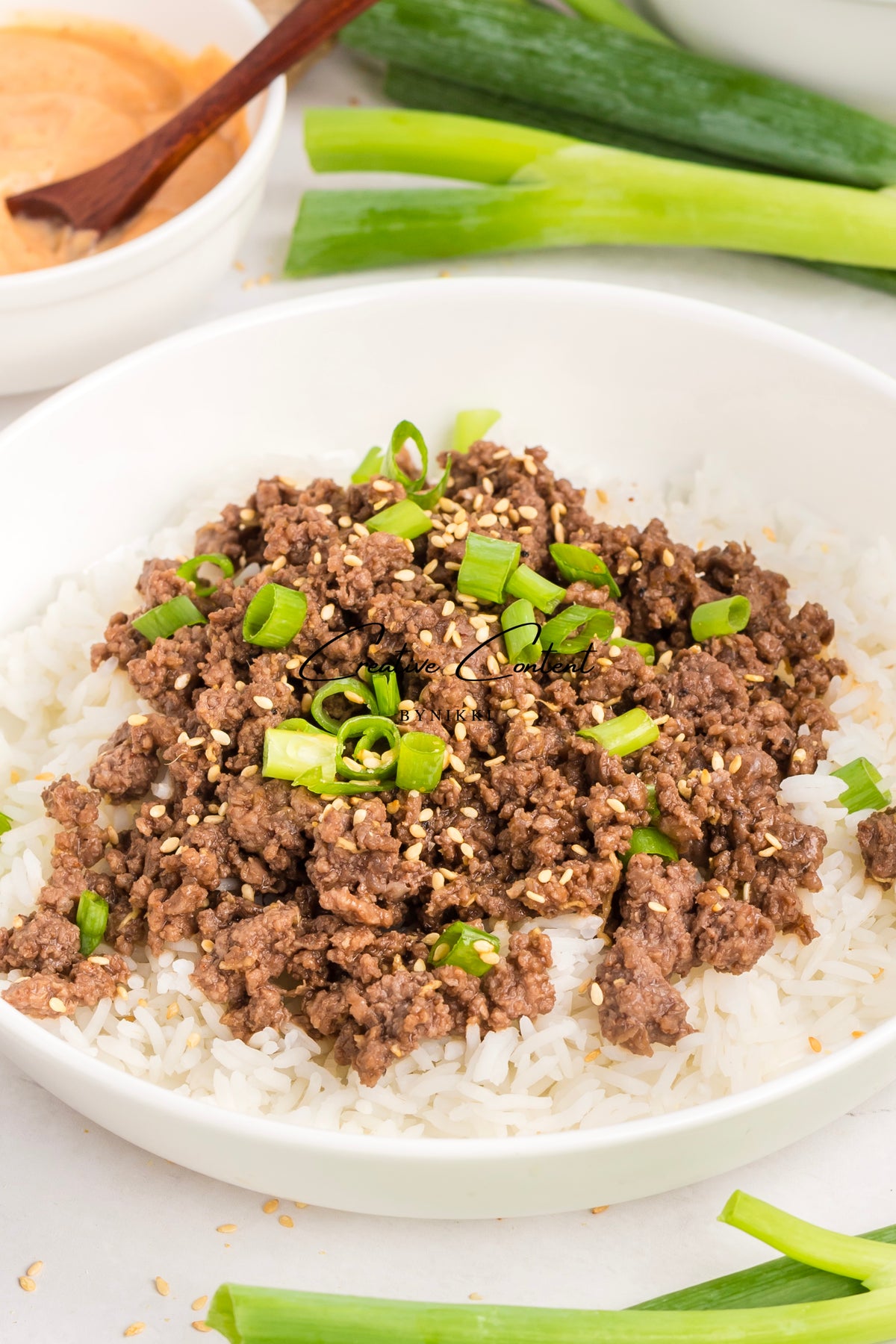 A bowl of Korean Beef Bulgogi - Exclusive, featuring ground beef with gochujang sauce topped with sesame seeds and chopped green onions over white rice, with a side of dipping sauce and green onions nearby.