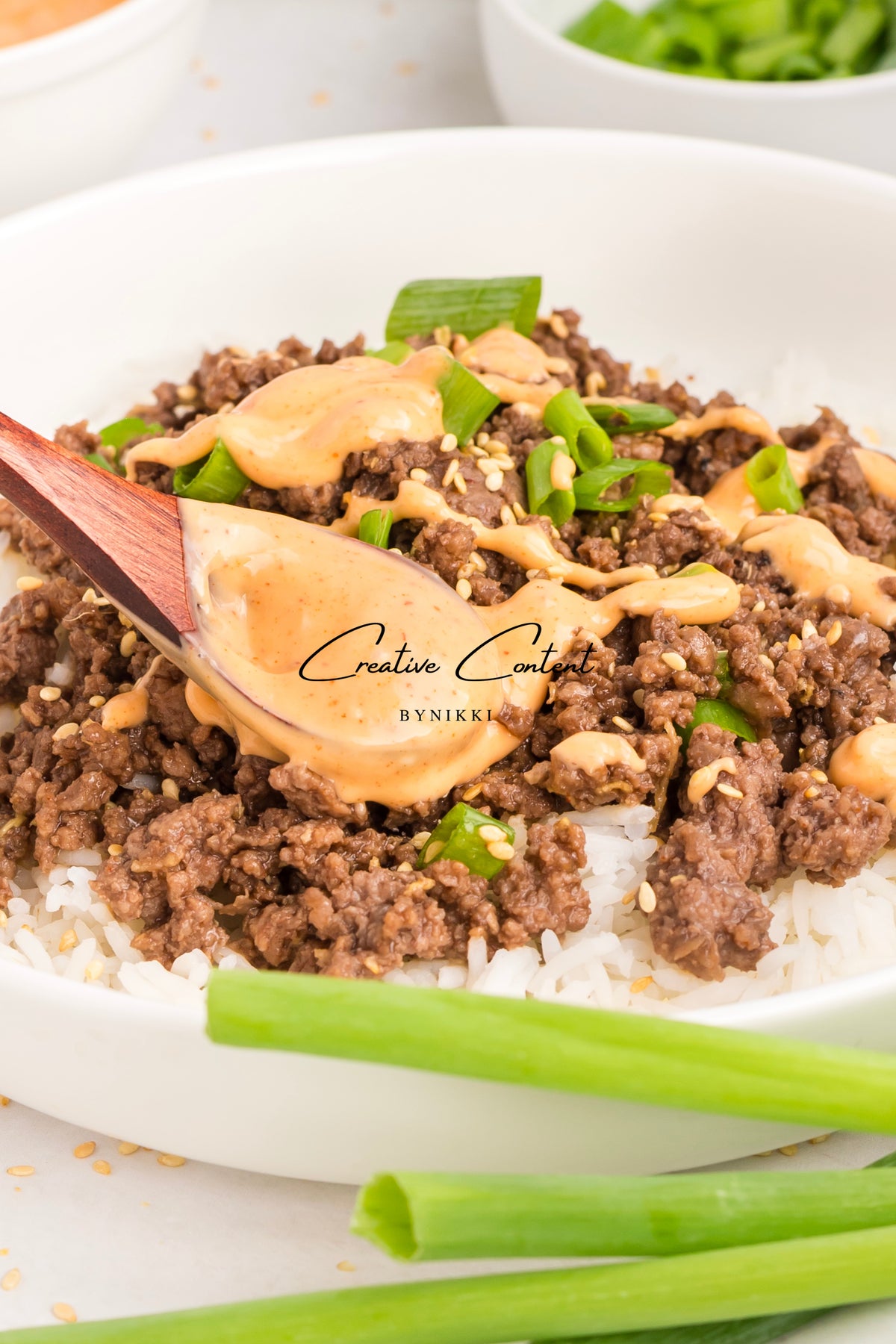 A wooden spoon scoops Korean Beef Bulgogi - Exclusive over white rice in a bowl, drizzled with gochujang sauce and garnished with sesame seeds and sliced green onions.