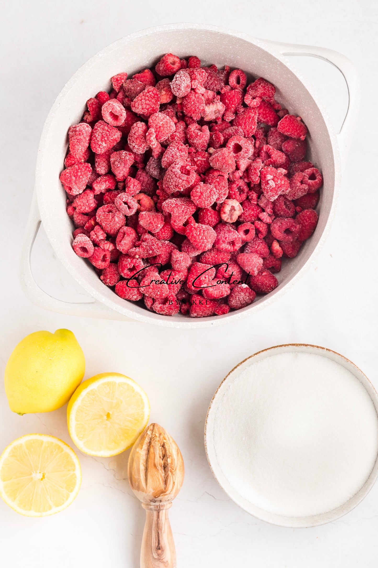 Frozen raspberries in a white bowl with lemons and a small bowl of sugar on a light background.