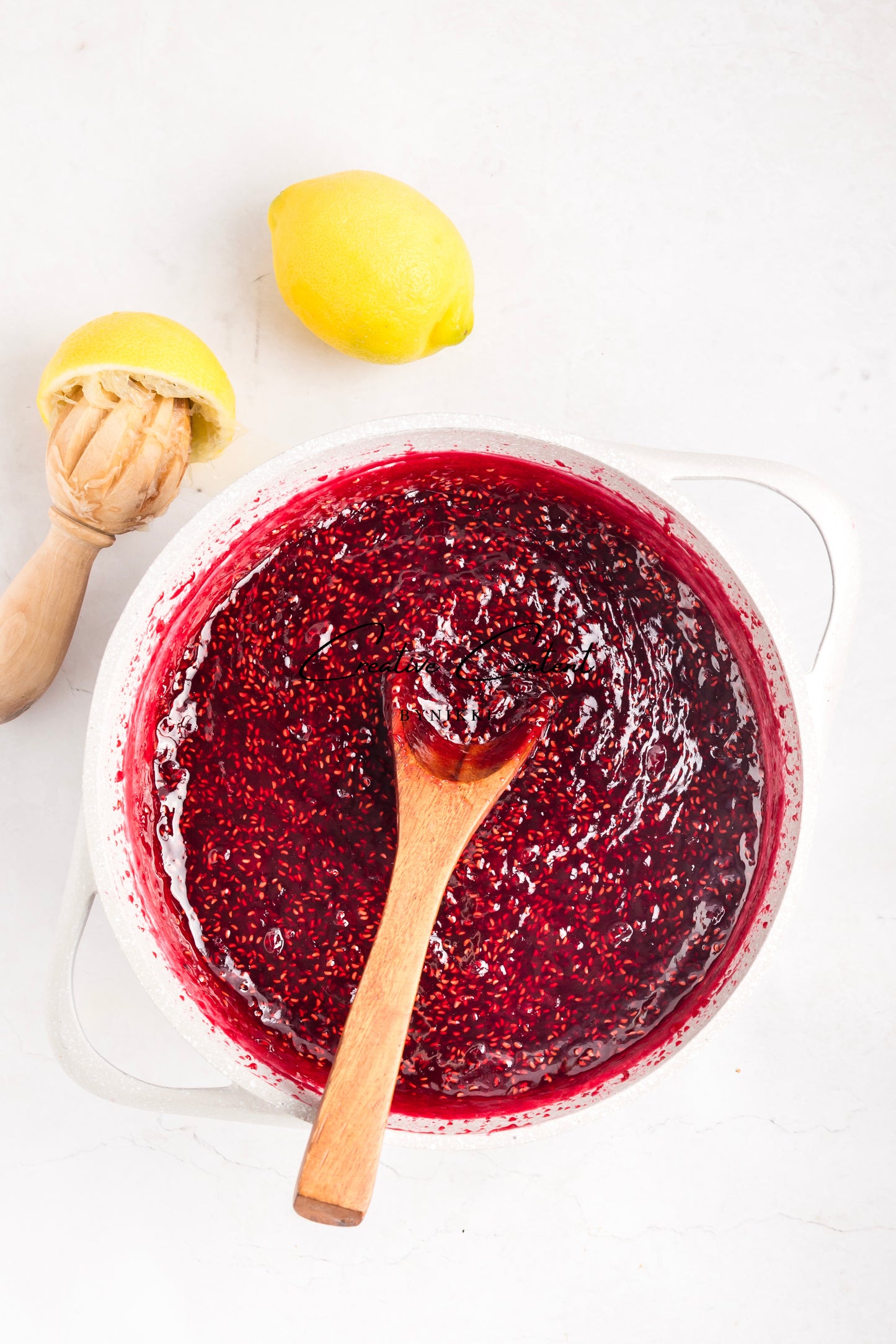 Red jam in a glass bowl with a wooden spoon, lemons, and a lemon squeezer on a white background