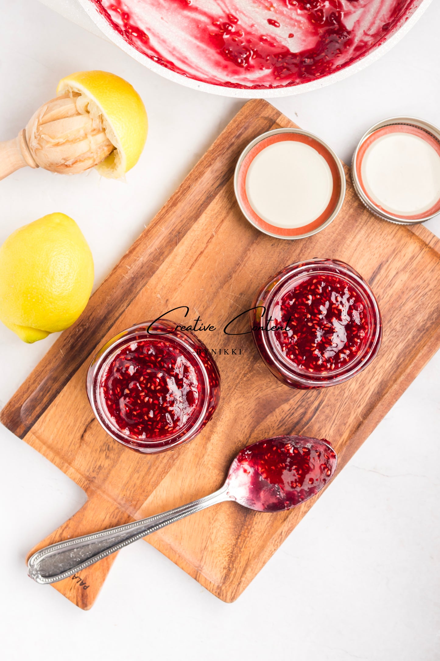 Two jars of red jam on a wooden cutting board with lemons and a bowl of jam in the background.