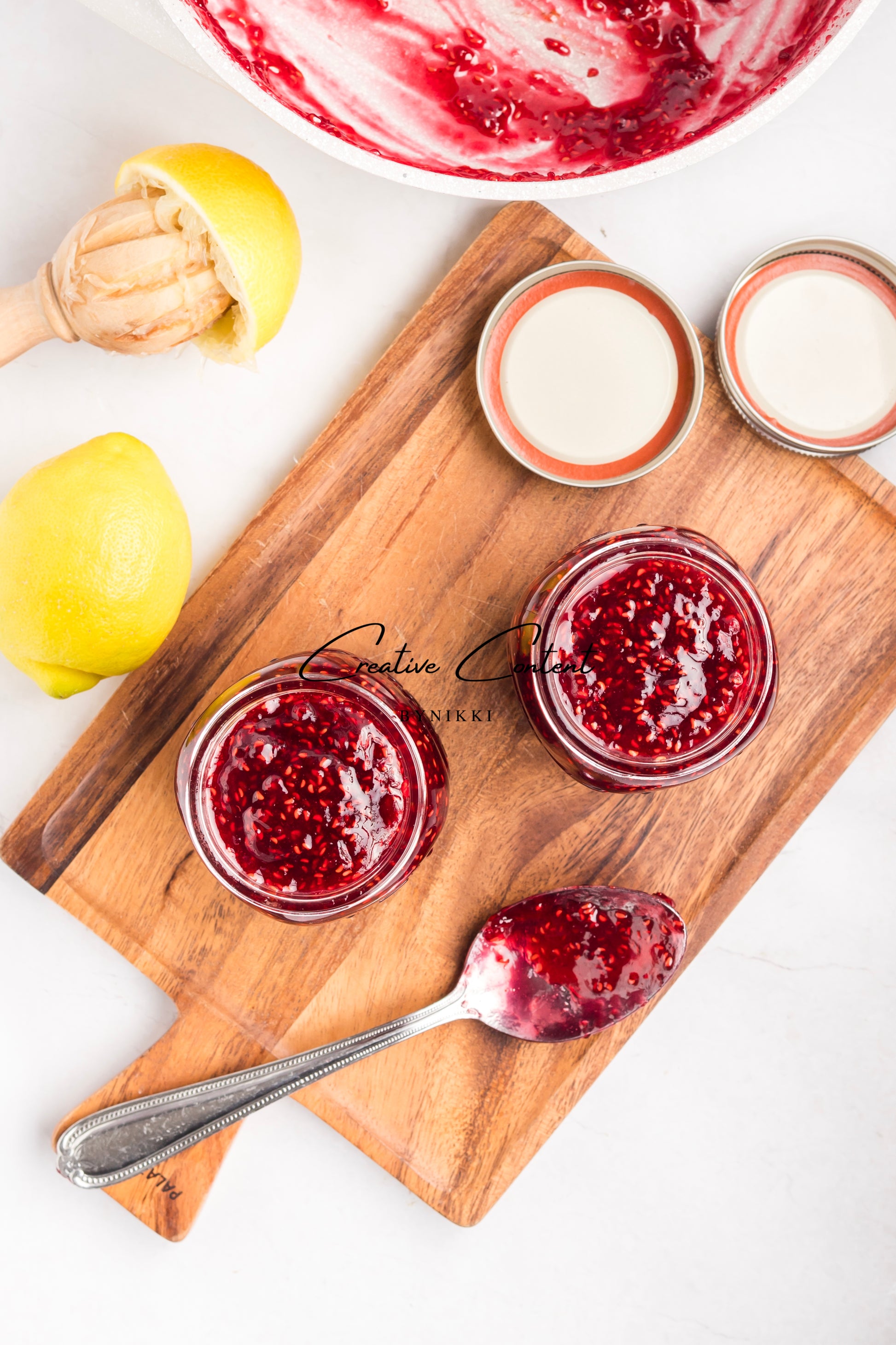 Two jars of red jam on a wooden cutting board with lemons and a bowl of jam in the background.
