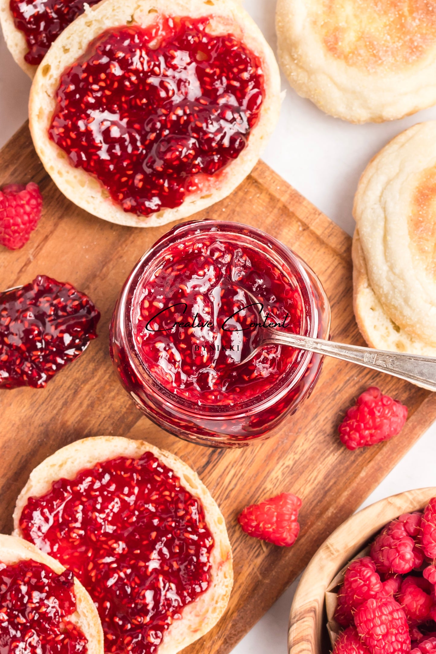 Glass jar of raspberry jam with a spoon, surrounded by English muffins and raspberries on a wooden board.