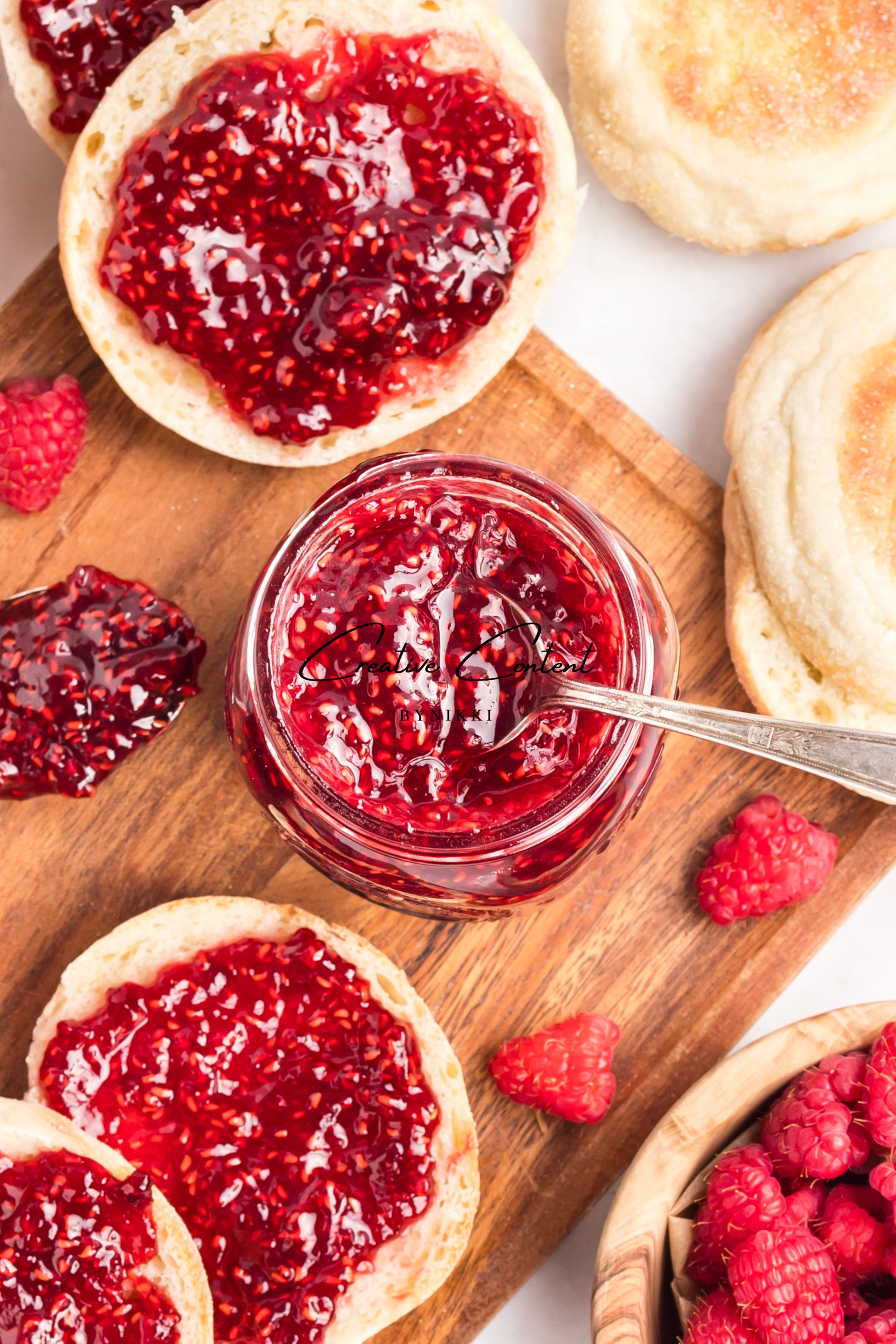 Glass jar of raspberry jam with a spoon, surrounded by English muffins and raspberries on a wooden board.