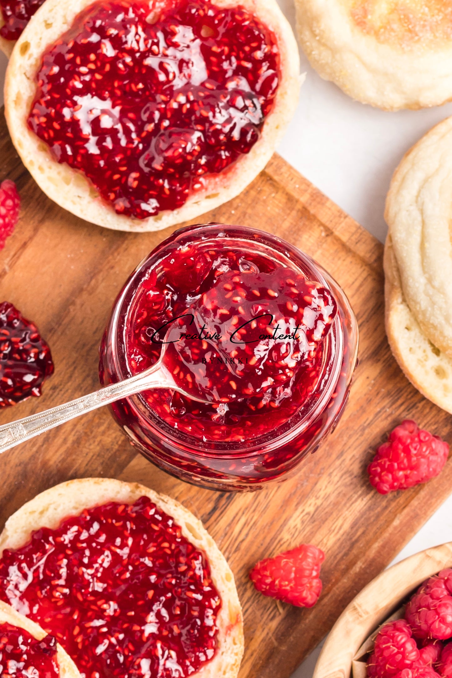 Jar of raspberry jam with a spoon, surrounded by English muffins and raspberries on a wooden board.