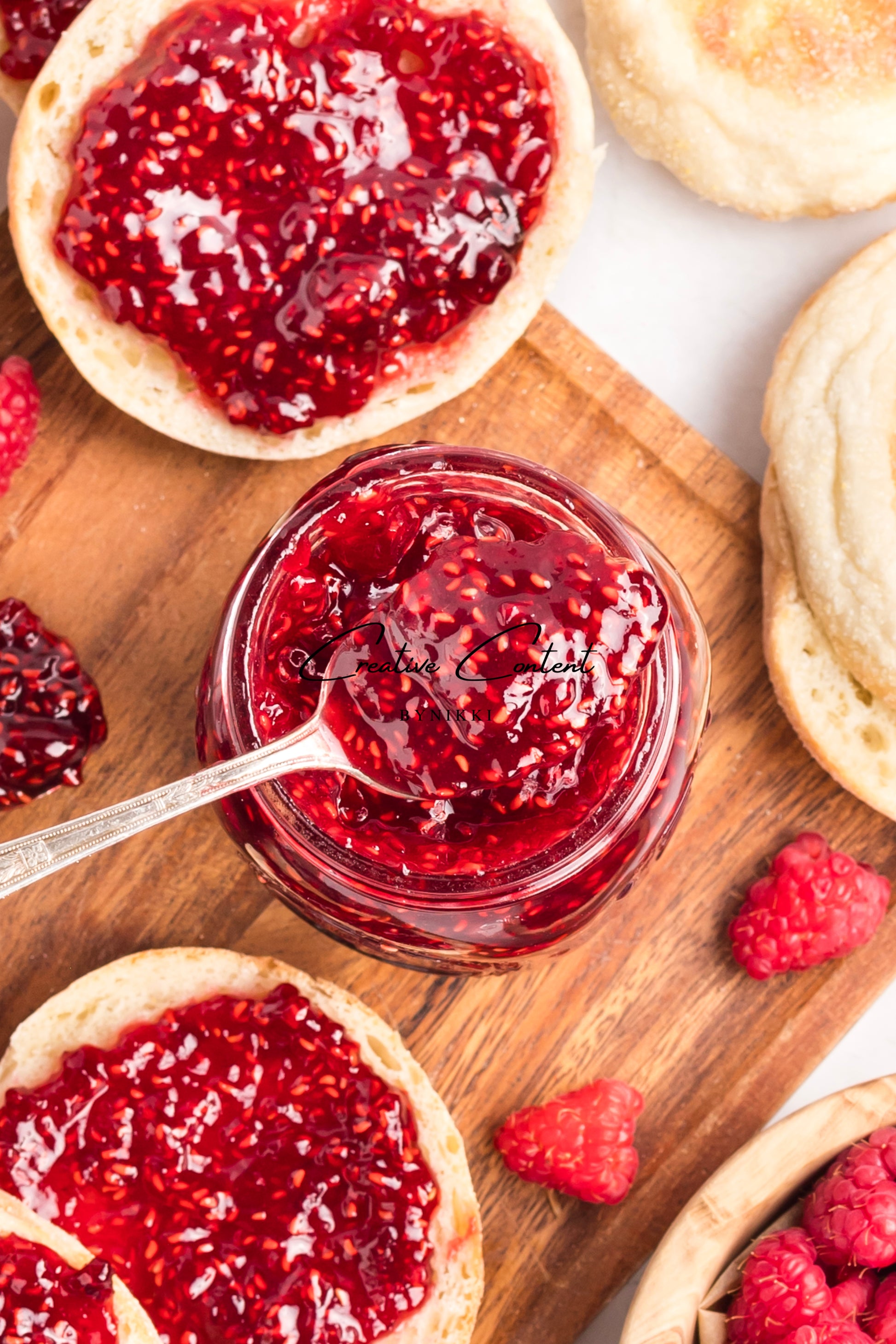 Jar of raspberry jam with a spoon, surrounded by English muffins and raspberries on a wooden board.