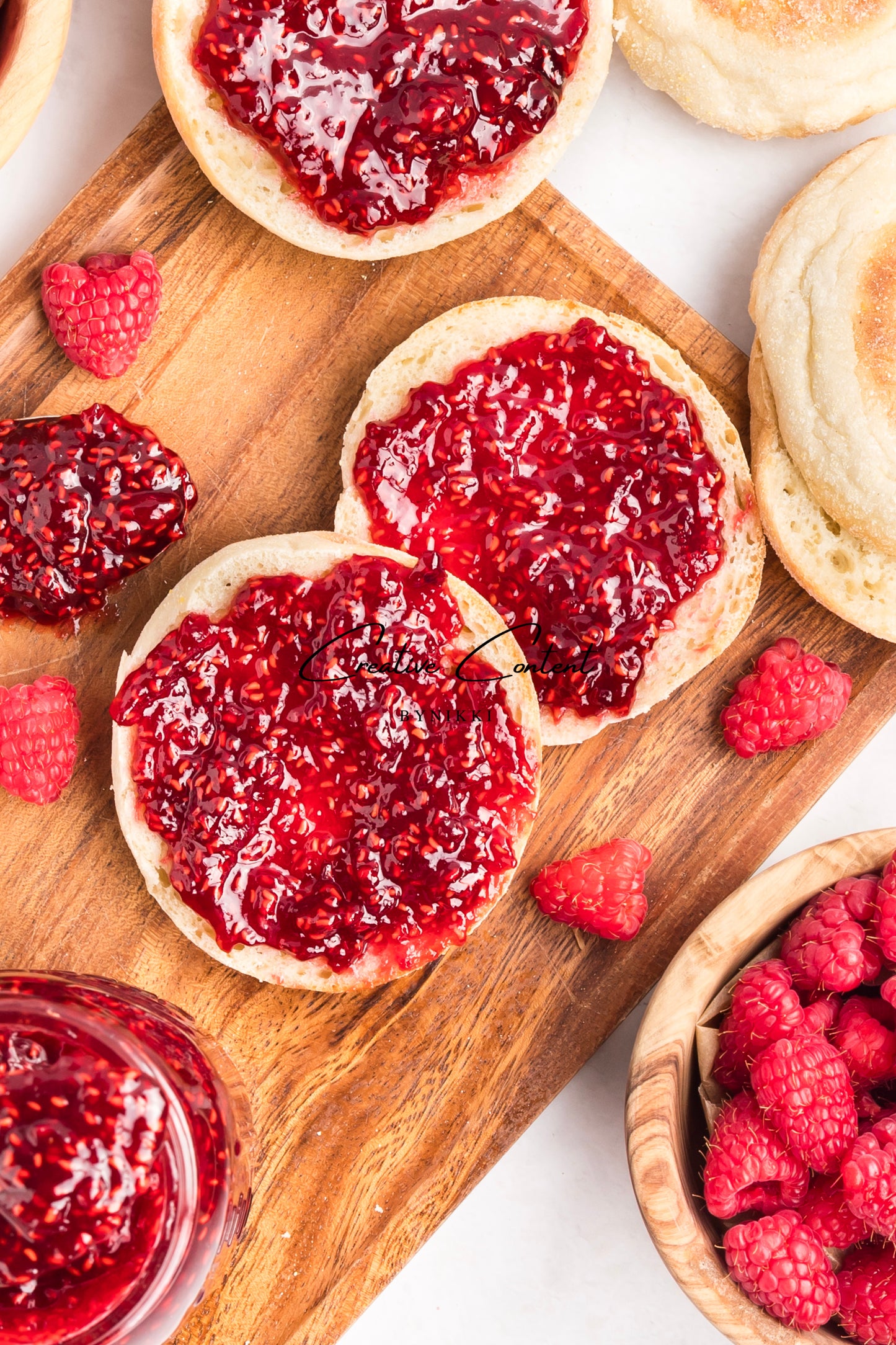 English muffins with raspberry jam on a wooden board, surrounded by raspberries.