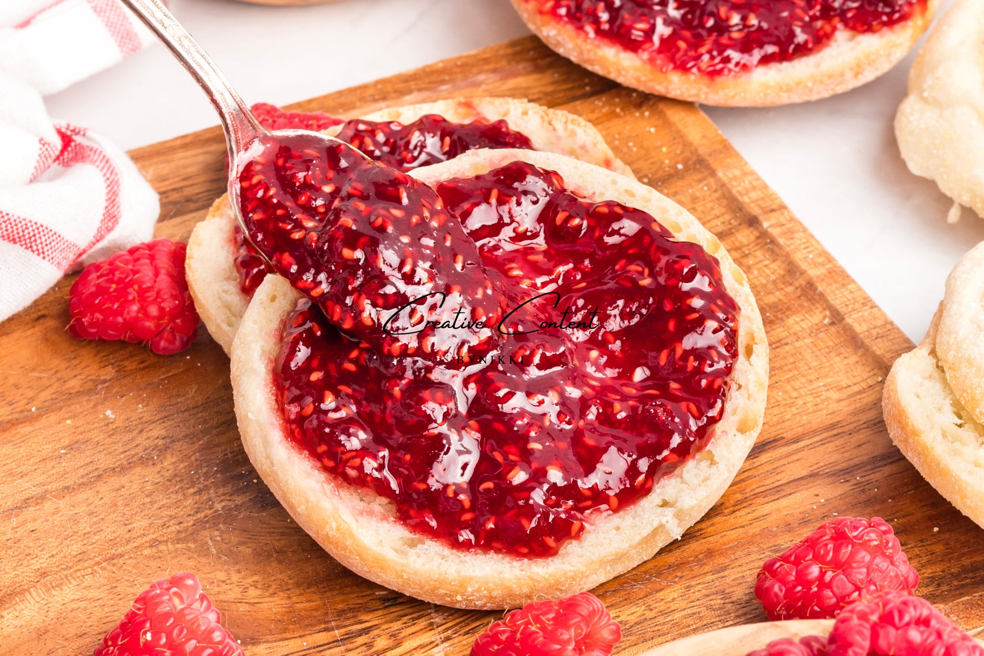 English muffin with raspberry jam on a wooden cutting board with raspberries around.