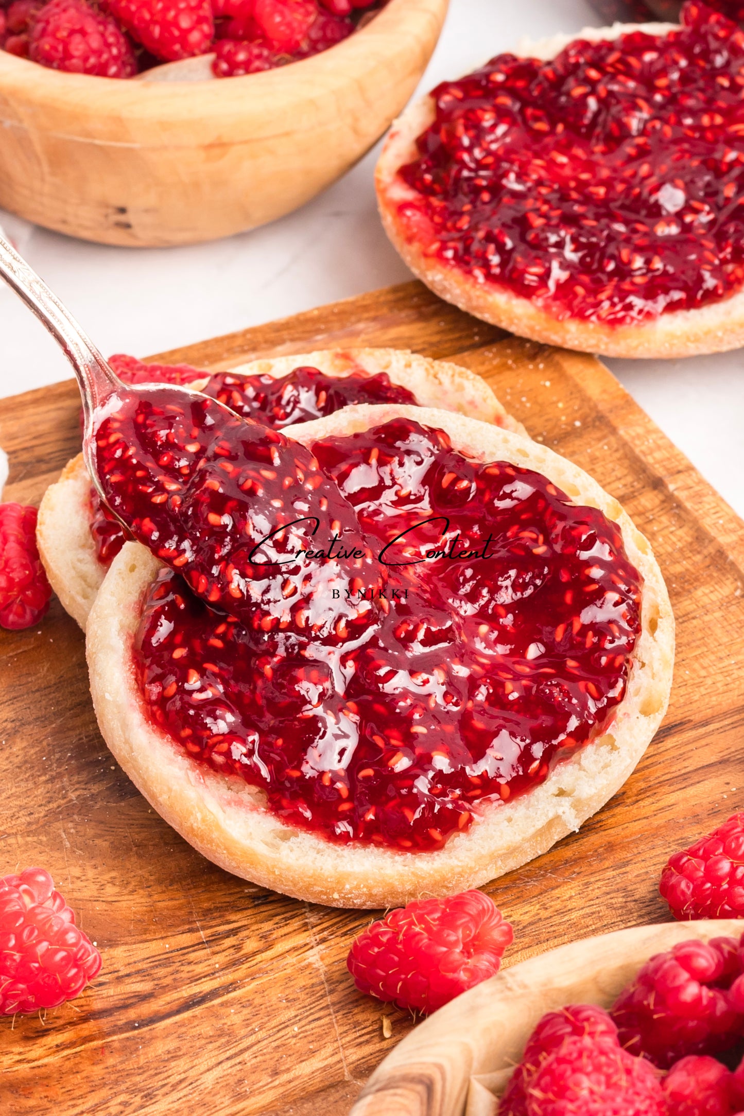 Raspberry jam spread on a biscuit with a spoon, surrounded by fresh raspberries on a wooden board.