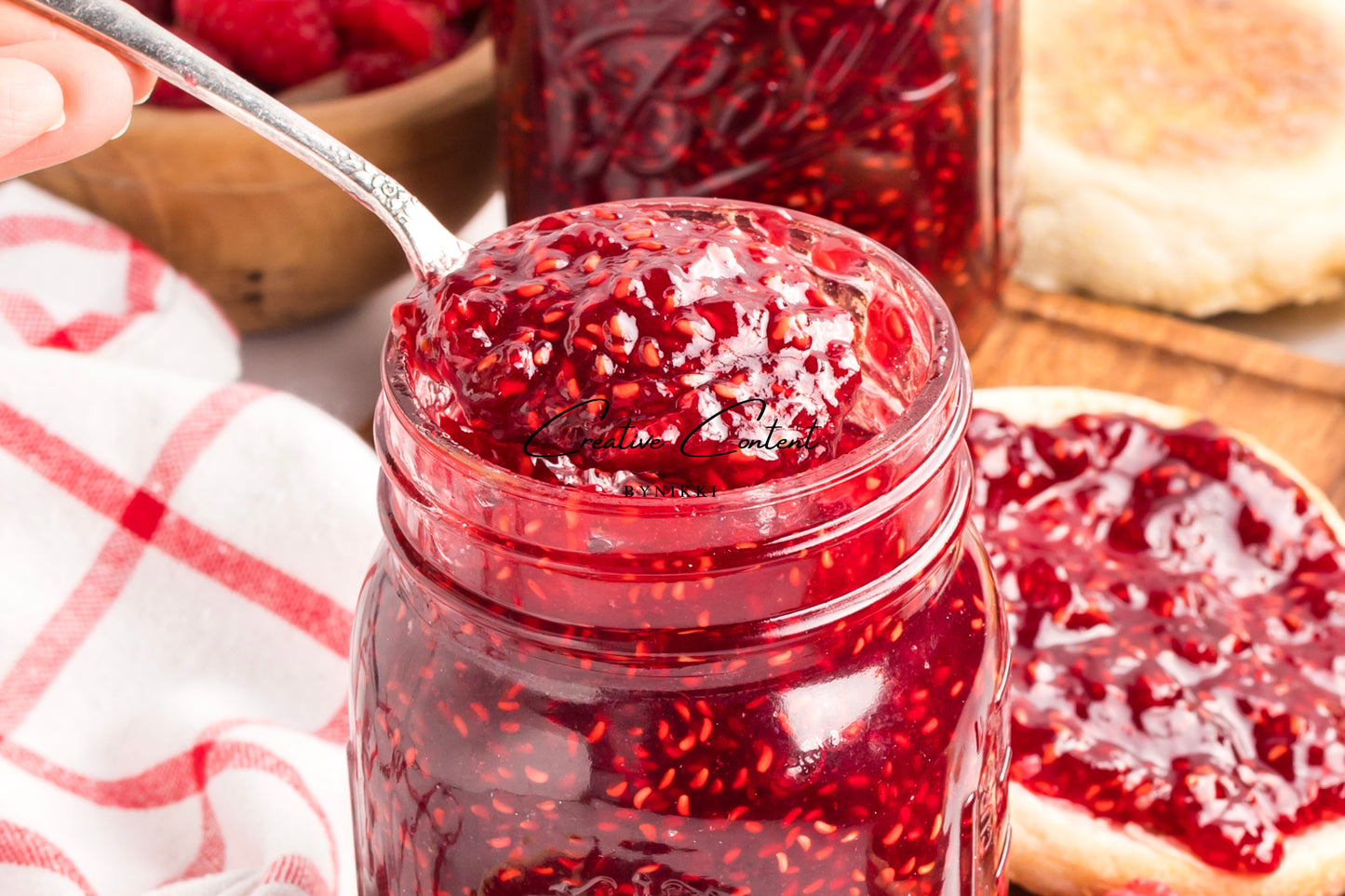 Jar of red raspberry jam with a spoonful being taken, surrounded by a checkered cloth and bread.