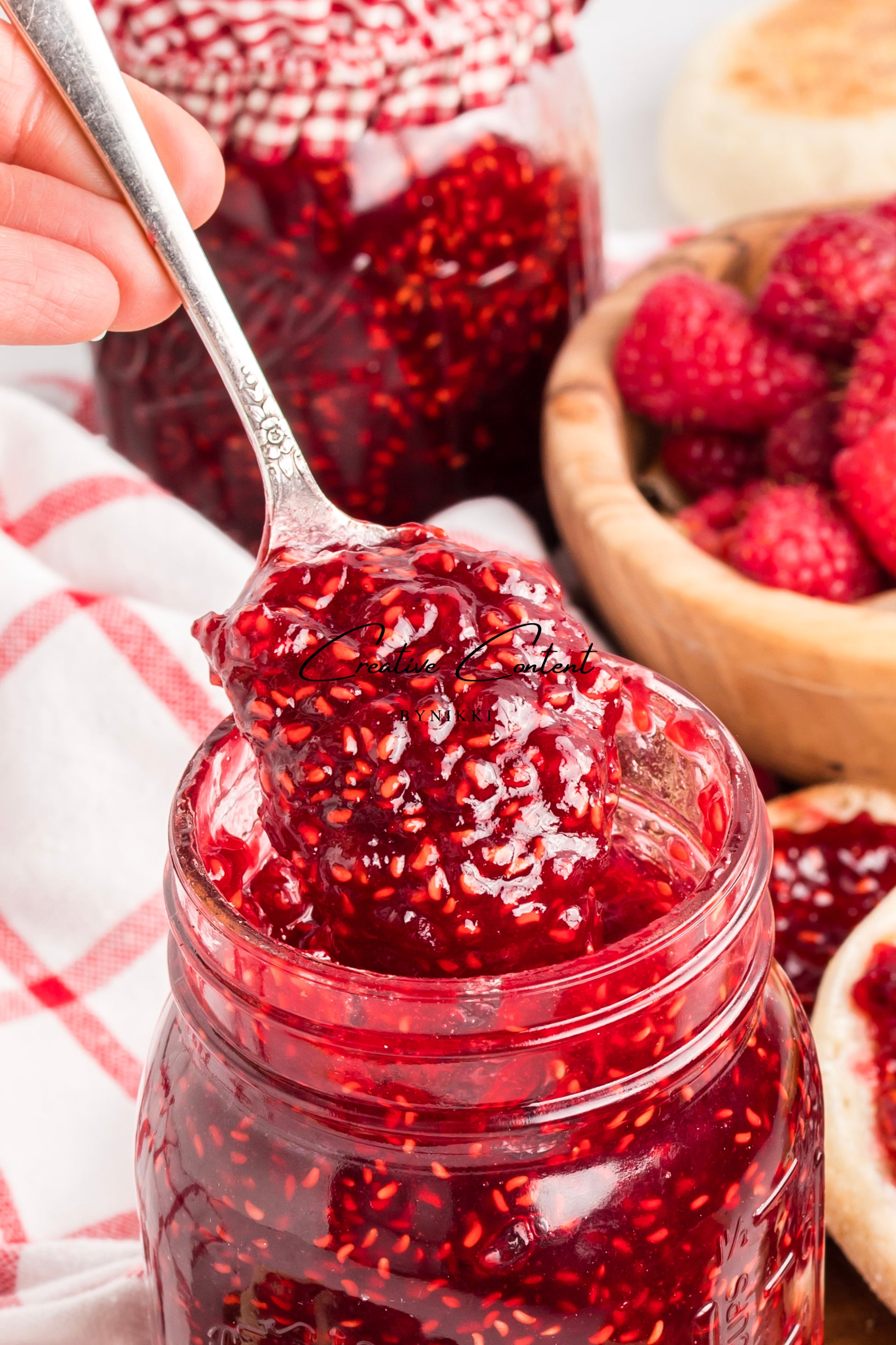 Jar of red raspberry jam with a spoonful being lifted, surrounded by fresh raspberries.